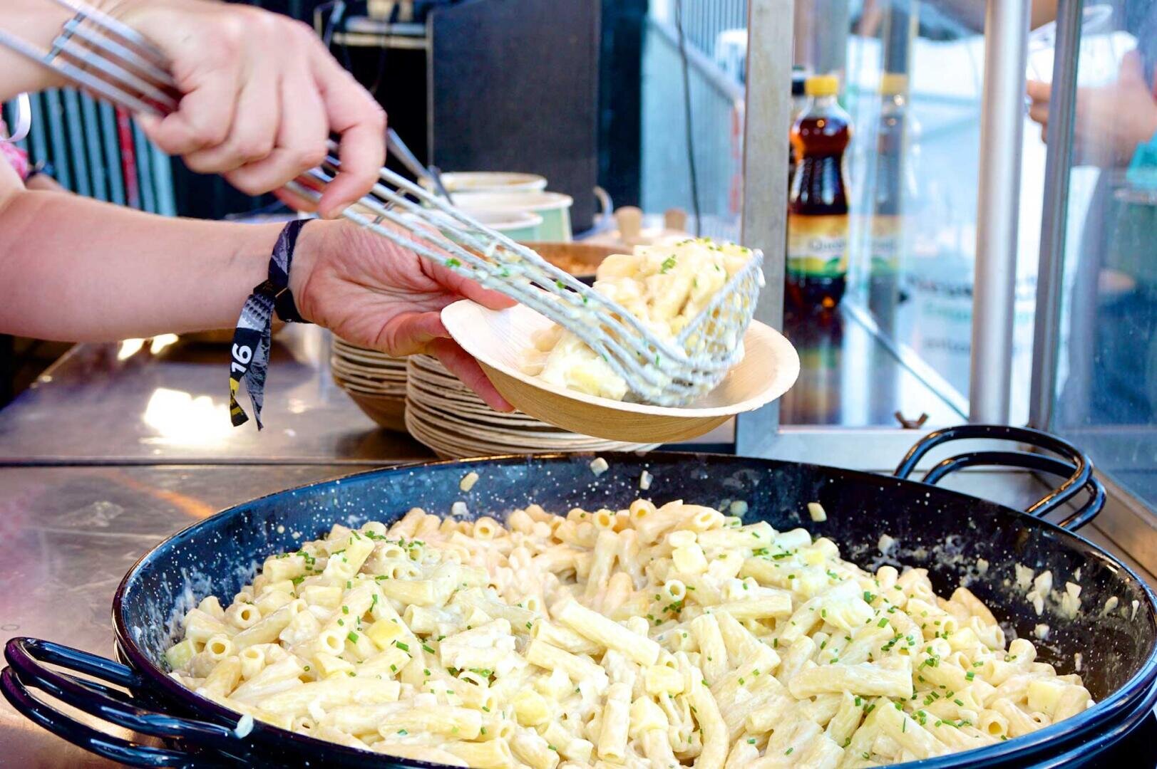 Eine Person serviert cremige Penne-Nudeln mit Kräutern aus einer großen schwarzen Pfanne mit einer Zange in eine Schüssel. Im Hintergrund sind Stapel von Schüsseln und Flaschen zu sehen.