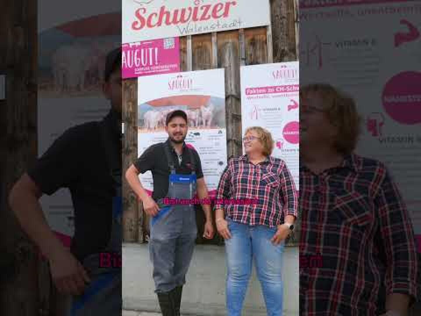 Ein Mann in Latzhose und Stiefeln steht neben einer Frau in einem karierten Hemd. Sie stehen lächelnd vor einem Gebäude mit Plakaten und einem Schild mit der Aufschrift "Schwizer Wilerstadt".