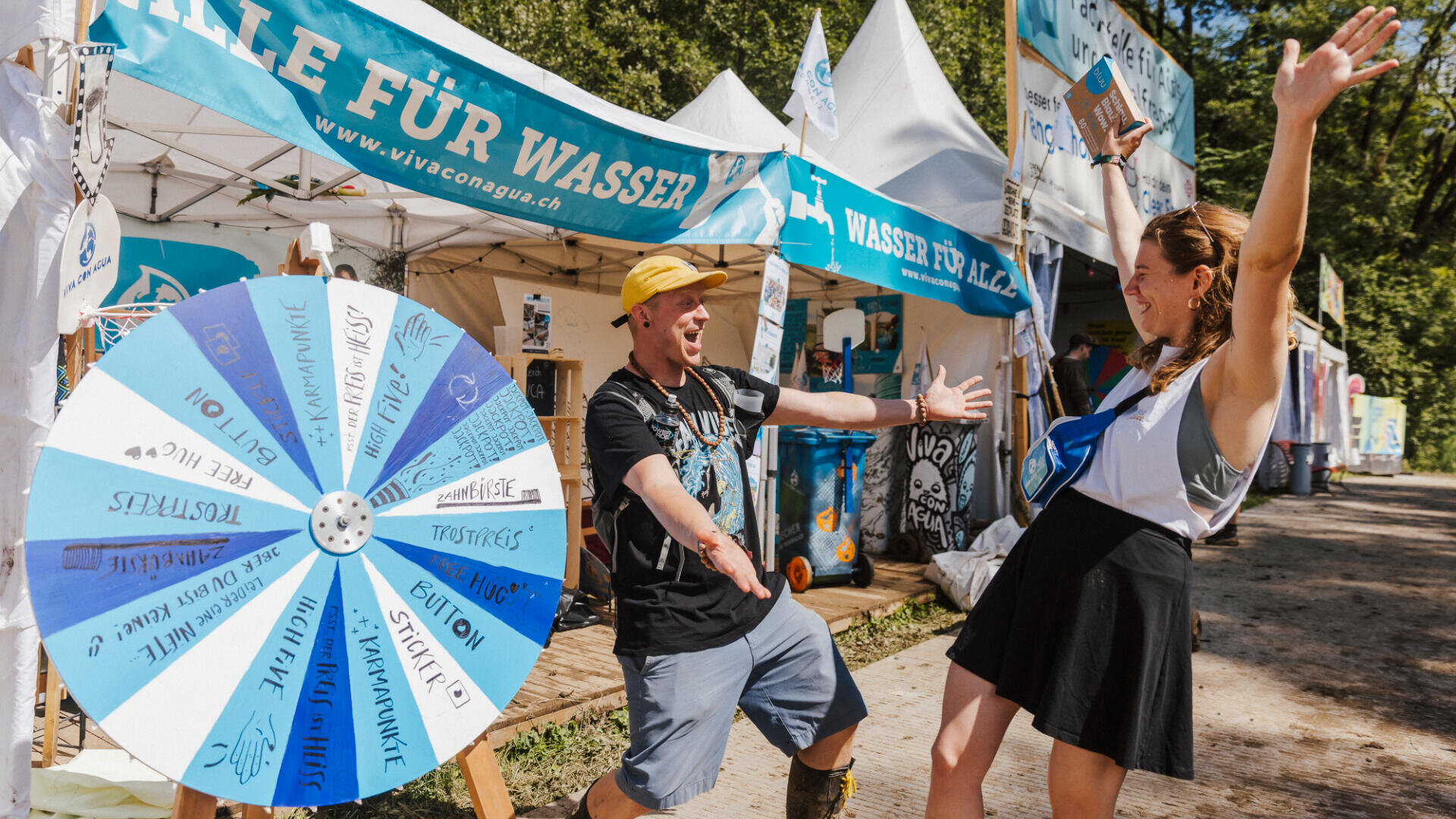 Zwei Personen feiern vor einem Glücksrad" an einem Stand, der auf einem Outdoor-Festival für sauberes Wasser wirbt, mit Bannern, Zelten und Grünpflanzen im Hintergrund.