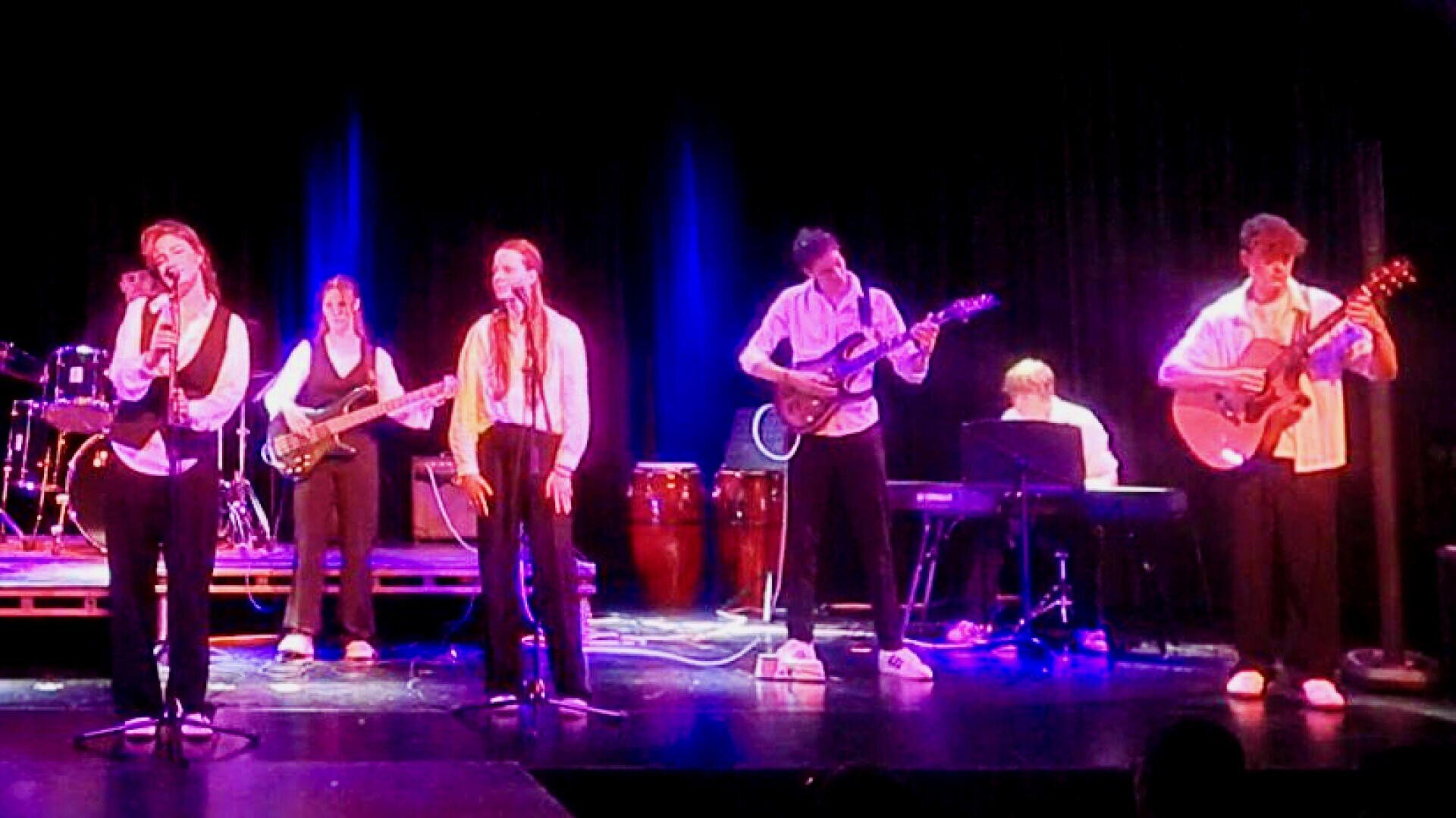 Auf der Bühne tritt eine sechsköpfige Band auf, darunter zwei Sänger mit Mikrofonen, ein Bassist, zwei Gitarristen und ein Keyboarder, während im Hintergrund eine bunte Bühnenbeleuchtung und ein Schlagzeug zu sehen sind.