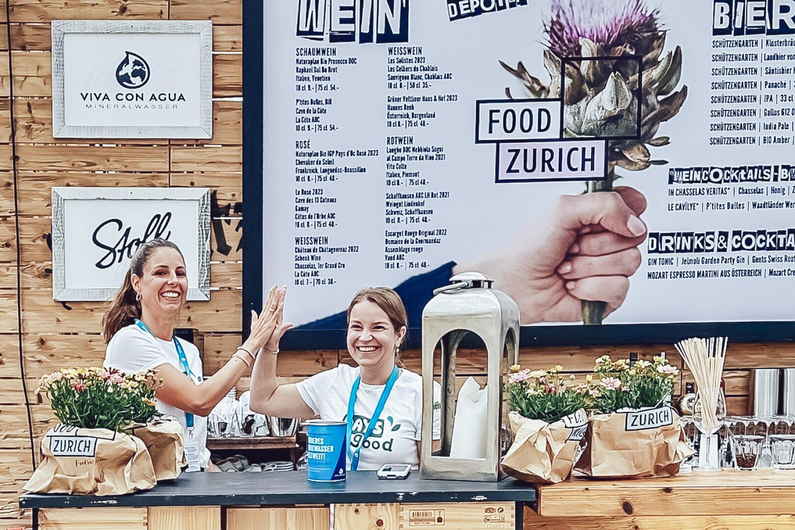 Zwei Frauen stehen lächelnd und sich zuprostend hinter einer mit Blumen und Papiertüten dekorierten Theke mit der Aufschrift Zürich. Im Hintergrund sind eine große Menütafel und Dekorationsartikel bei einer Veranstaltung im Freien zu sehen.