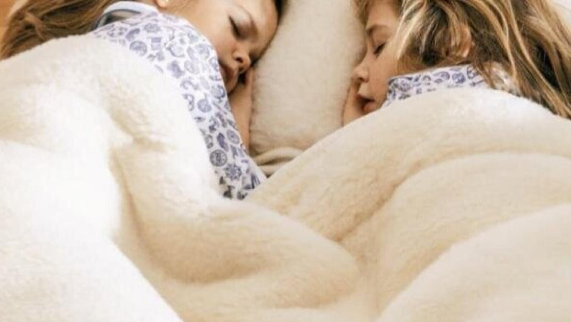 Zwei kleine Kinder, die gemusterte Schlafanzüge tragen, schlafen friedlich nebeneinander unter einer flauschigen weißen Decke. Ihre Gesichter liegen dicht beieinander, und sie scheinen es bequem und gemütlich zu haben.