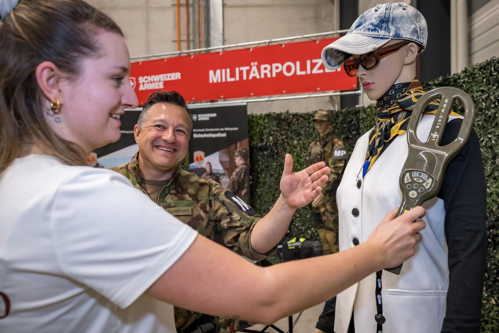 Eine Frau benutzt einen handgehaltenen Metalldetektor an einer Schaufensterpuppe, während ein Mann in Schweizer Militäruniform lächelnd zusieht; im Hintergrund ist ein Schild der Militärpolizei zu sehen.