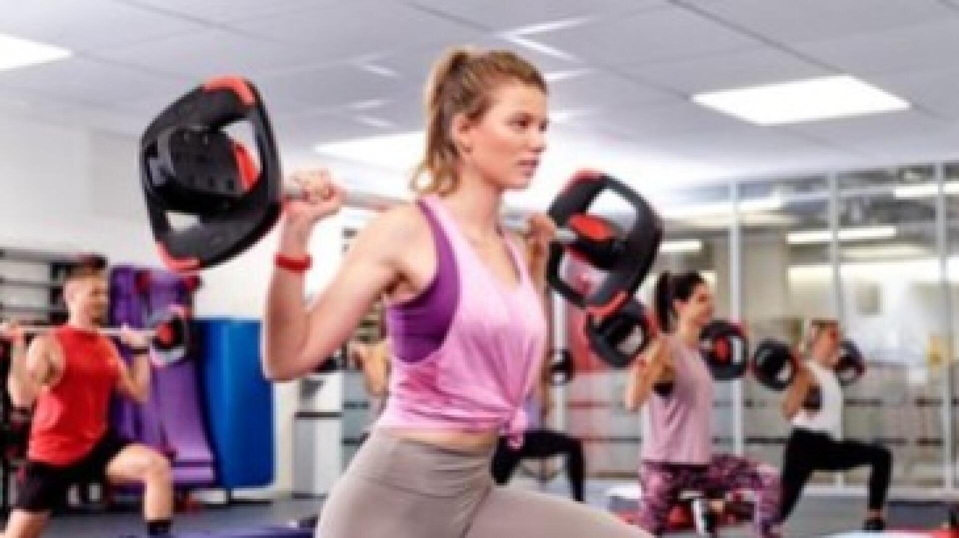 Ein Gruppenfitnesskurs, der in einer hellen Turnhalle Ausfallschritte mit Hanteln macht; eine Frau im Vordergrund trägt ein rosa Oberteil und graue Leggings, während andere im Hintergrund trainieren.