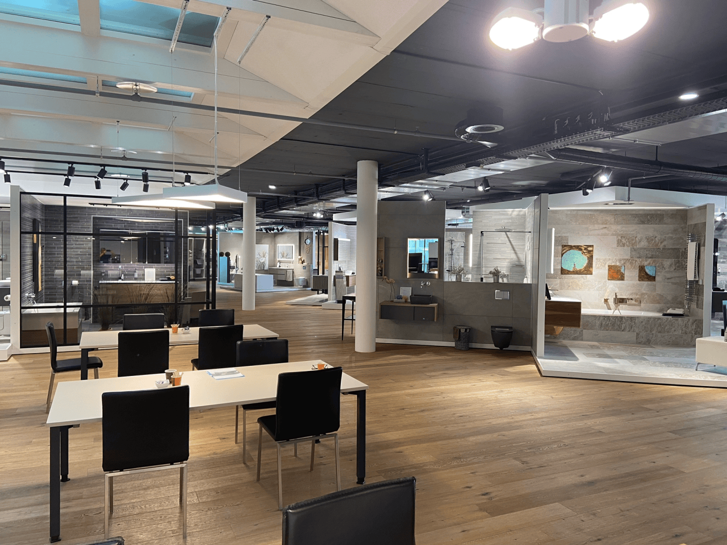 Ein moderner, offen gestalteter Ausstellungsraum mit Holzböden, Ausstellungsflächen mit Badarmaturen und mehreren schwarzen Stühlen und hellen Tischen im Vordergrund. Der Raum ist gut beleuchtet und geordnet.