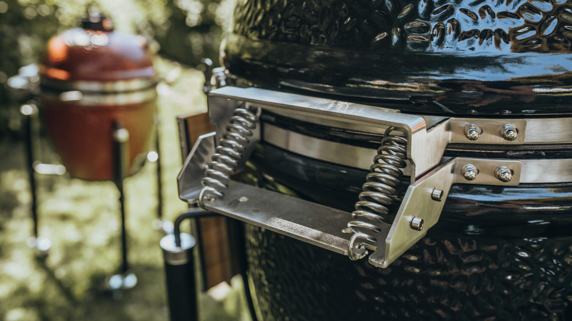 Nahaufnahme eines schwarzen Grillgriffs aus Keramik mit Metallfedern im Freien, im Hintergrund ist ein weiterer, ähnlicher roter Grill zwischen Gras und Bäumen verschwommen zu sehen.