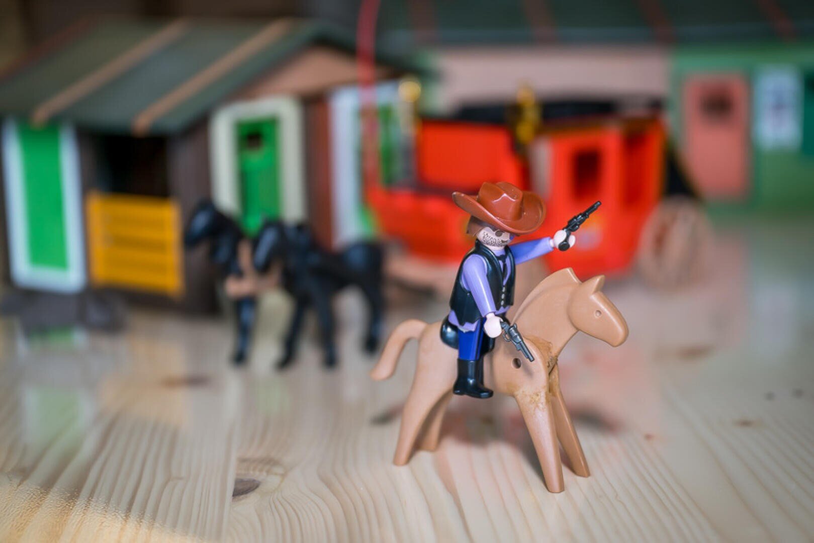 Eine Playmobil-Cowboy-Figur mit Hut und zwei Pistolen reitet auf einem Spielzeugpferd auf einer hölzernen Fläche, wobei Spielzeugpferde, eine Kutsche und ein Haus im Hintergrund verschwimmen.