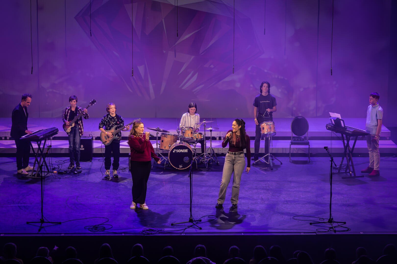 Eine siebenköpfige Band spielt auf der Bühne mit Keyboards, Gitarren, Schlagzeug und Percussion. Zwei Frauen stehen vorne und singen in Mikrofone. Hinter ihnen befinden sich eine violette Beleuchtung und ein geometrischer Bühnenhintergrund.