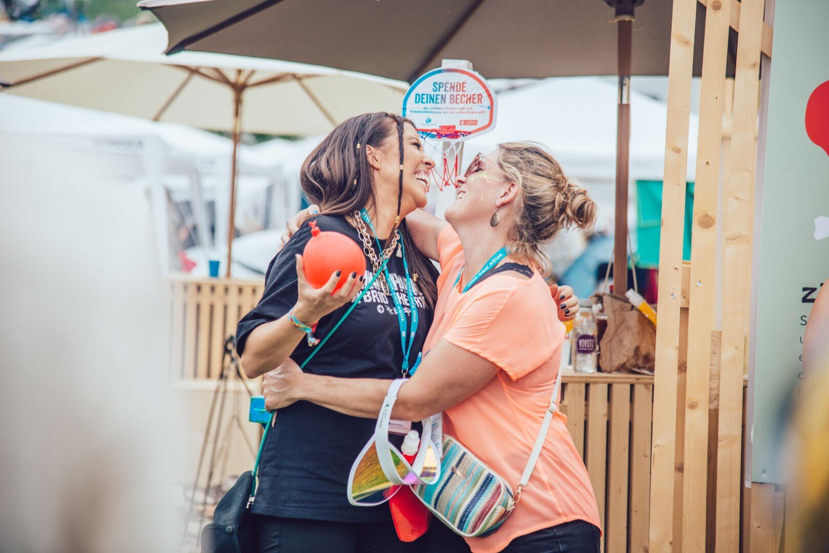 Zwei Frauen lächeln und umarmen sich im Freien bei einer Veranstaltung, eine hält einen roten Ball. Sie wirken glücklich und sind von Sonnenschirmen und Holzkonstruktionen umgeben, mit Menschen und Ständen im Hintergrund.