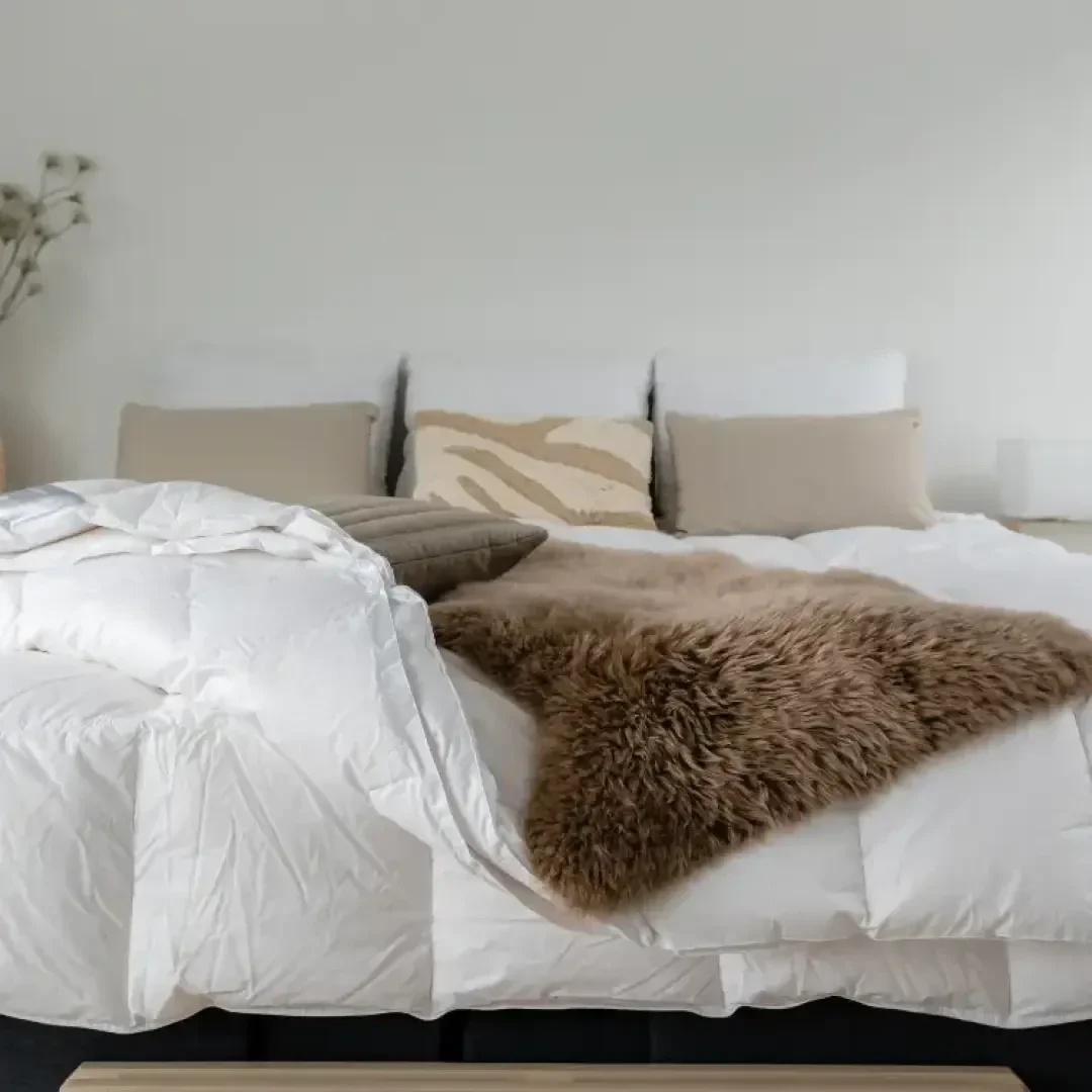 Ein gemütliches, ordentlich gemachtes Bett mit einer weißen Bettdecke, beigen und weißen Kissen und einer braunen Kunstfelldecke vor einer schlichten hellen Wand mit einer kleinen getrockneten Pflanze im Hintergrund.