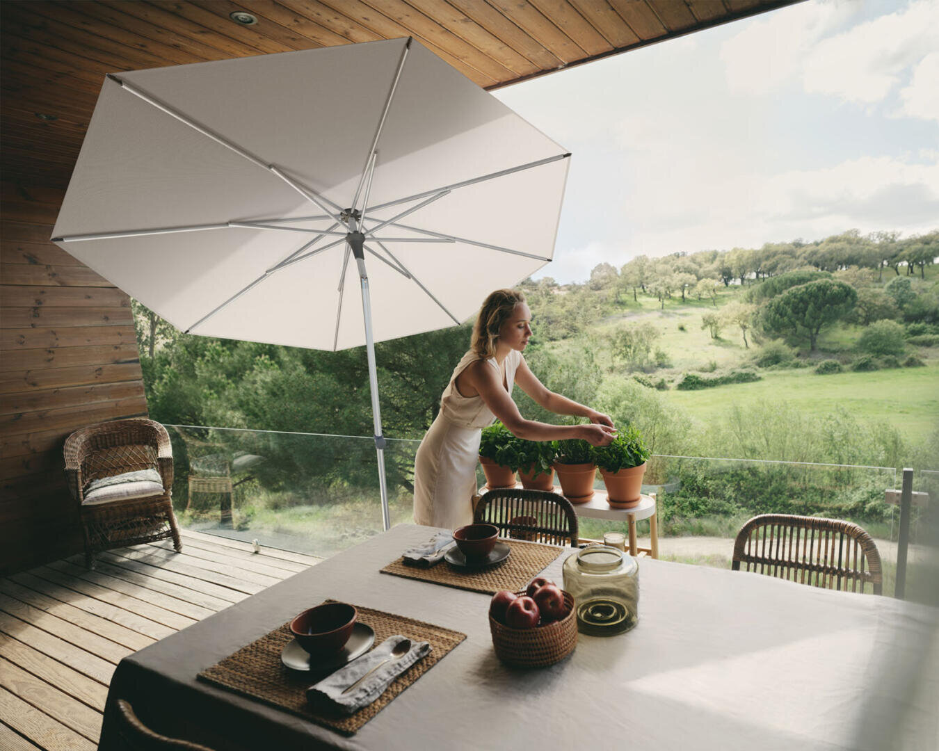 Eine Frau kümmert sich um Topfpflanzen auf einer Holzterrasse mit einem großen Sonnenschirm und Blick auf eine grüne Landschaft. Ein Tisch ist mit Tischsets, Schüsseln und einer Schale mit Äpfeln gedeckt. Korbstühle umgeben den Tisch.