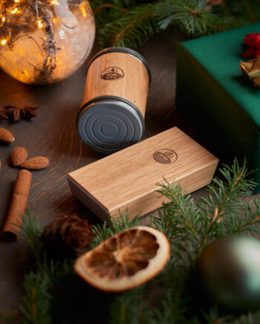 Eine festliche Szene mit einem Holzbehälter und einer rechteckigen Holzkiste, umgeben von immergrünen Zweigen, getrockneten Zitrusfrüchten, Gewürzen, einer grünen Geschenkbox und warmen Lichterketten.