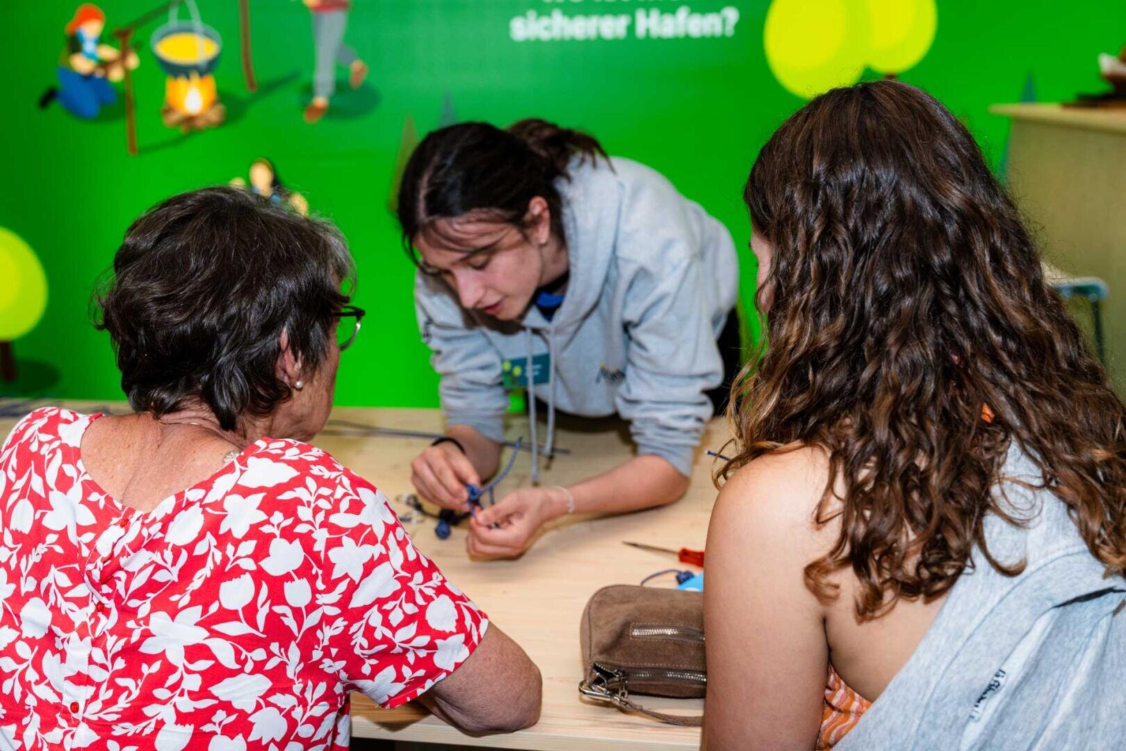 Drei Personen sitzen an einem Tisch und beschäftigen sich mit einer Bastelarbeit. Eine junge Person in einem grauen Kapuzenpulli hilft zwei anderen, einer mit kurzen grauen Haaren und geblümtem Hemd, der andere mit langen gewellten Haaren. Der Hintergrund ist hellgrün.