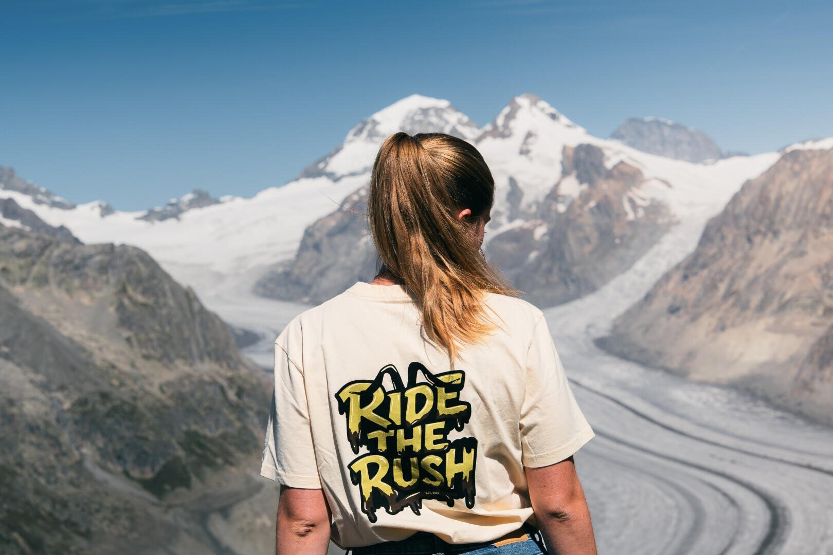 Eine Frau mit Pferdeschwanz, die ein T-Shirt mit der Aufschrift RIDE THE RUSH trägt, steht vor schneebedeckten Bergen und einem Gletscher unter strahlend blauem Himmel.
