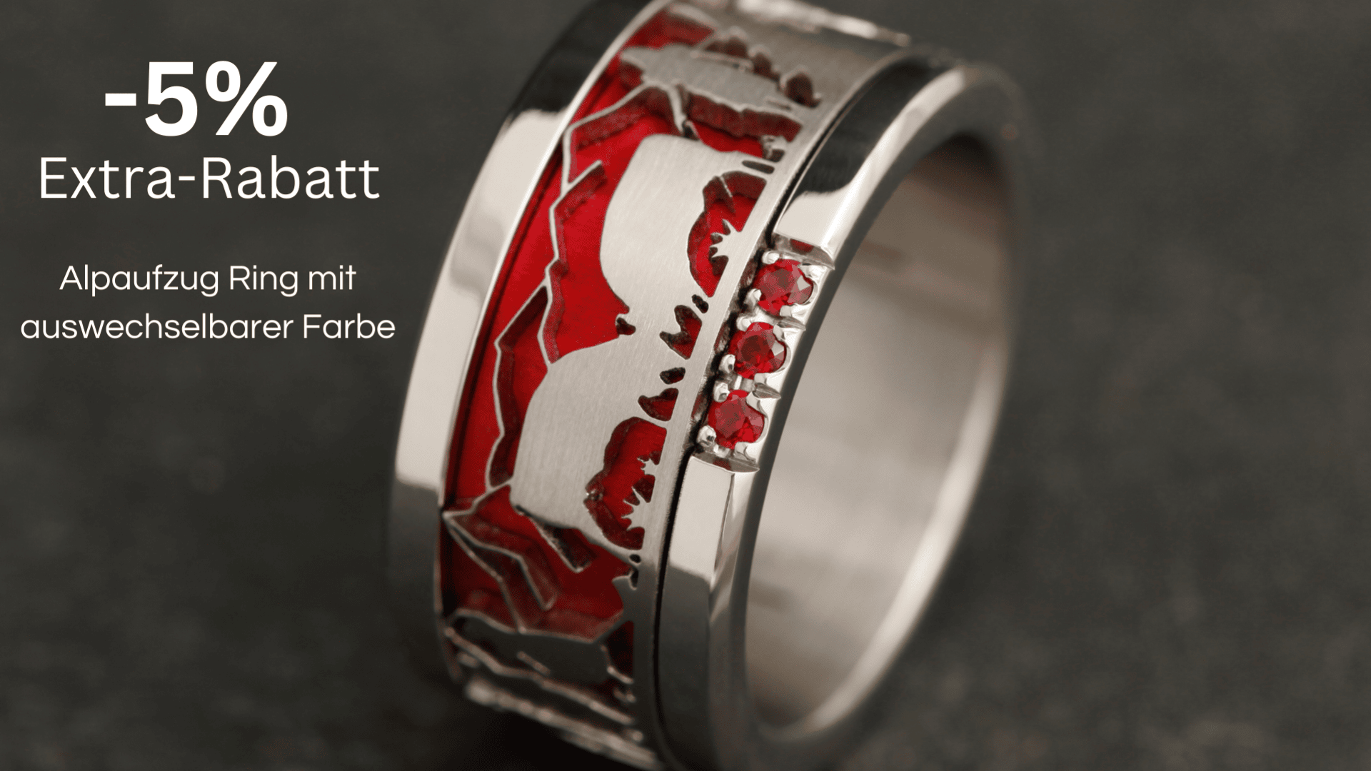Abgebildet ist ein silberner Ring mit einer roten, strukturierten Einlage und drei kleinen roten Edelsteinen. Der Text lautet: -5 % Extra-Rabatt. Alpaufzug-Ring mit austauschbarer Farbe.