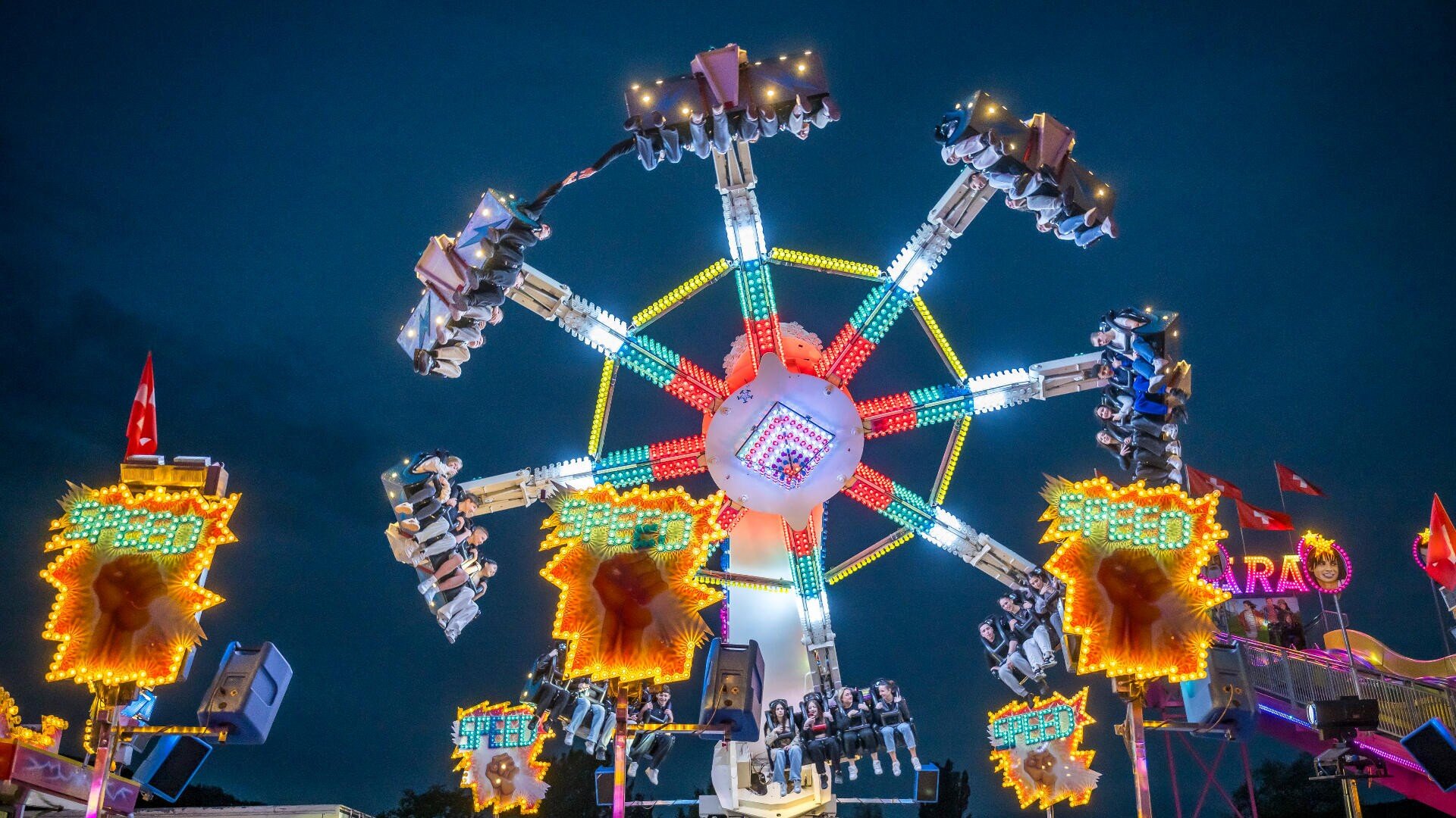Ein hell erleuchtetes Fahrgeschäft dreht sich nachts vor dem dunklen Himmel, mit Neonlichtern in Rot, Gelb, Blau und Grün. Die schwingenden Sitze sind erhöht, und bunte Schilder und Fahnen tragen zur festlichen Atmosphäre bei.