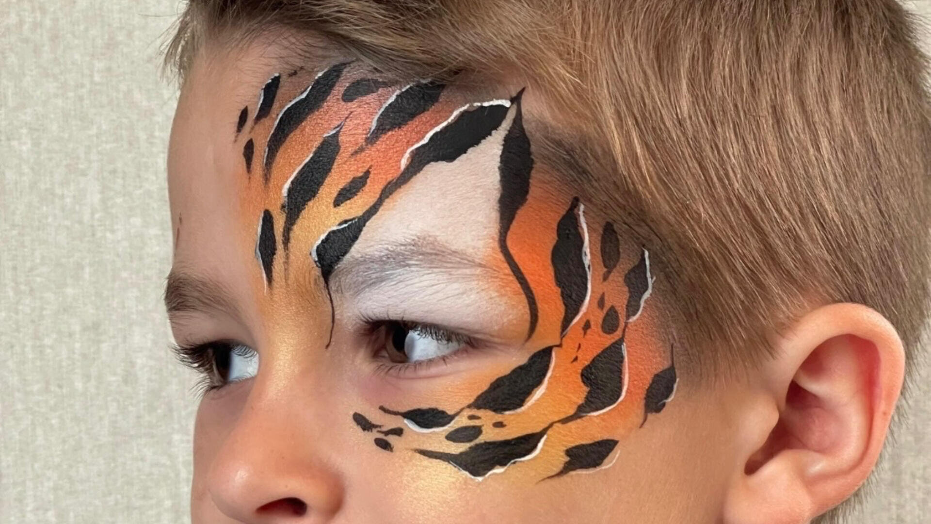 Nahaufnahme eines Kindergesichts mit detaillierter Tigerbemalung auf der Stirn und um die Augen, mit schwarzen Streifen, orangefarbenen und weißen Akzenten. Das Kind hat kurzes braunes Haar und blickt nach links.