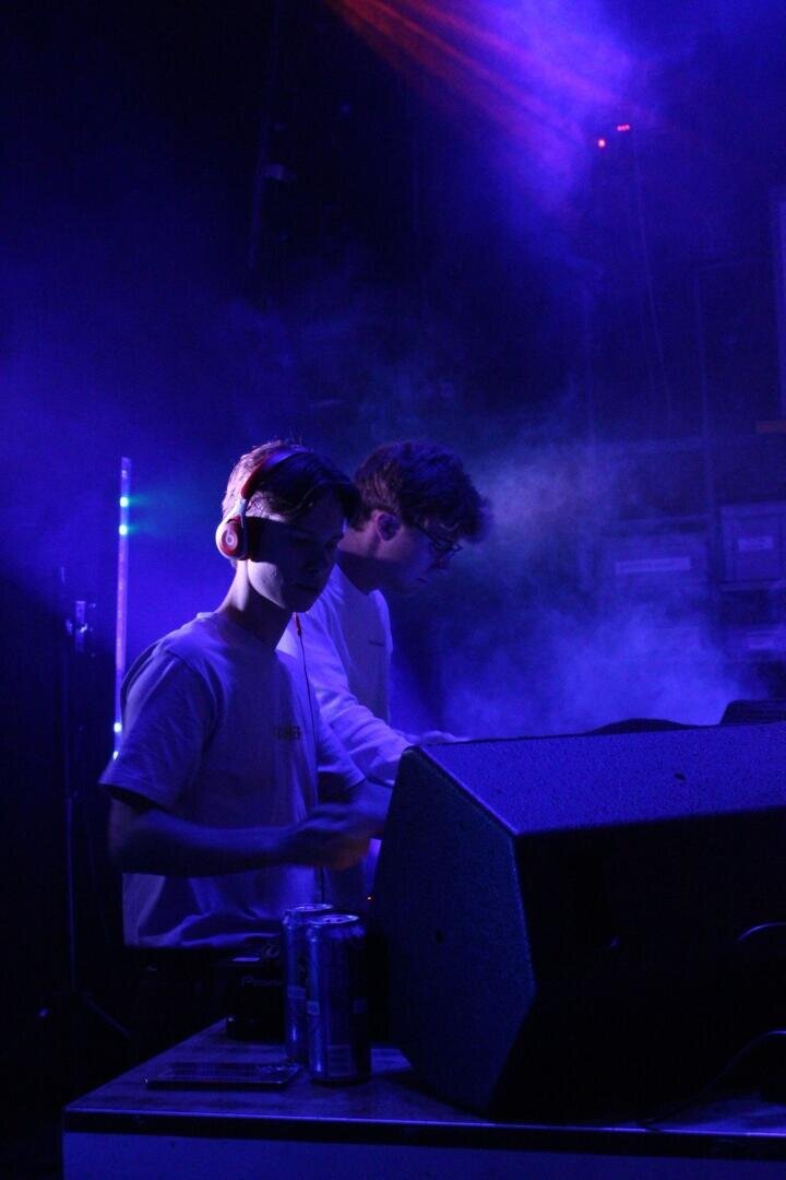 Zwei junge DJs in weißen Hemden stehen unter blauen und lila Lichtern auf der Bühne, umgeben von Nebel, im Vordergrund sind Tonanlagen und Energy-Drink-Dosen zu sehen.