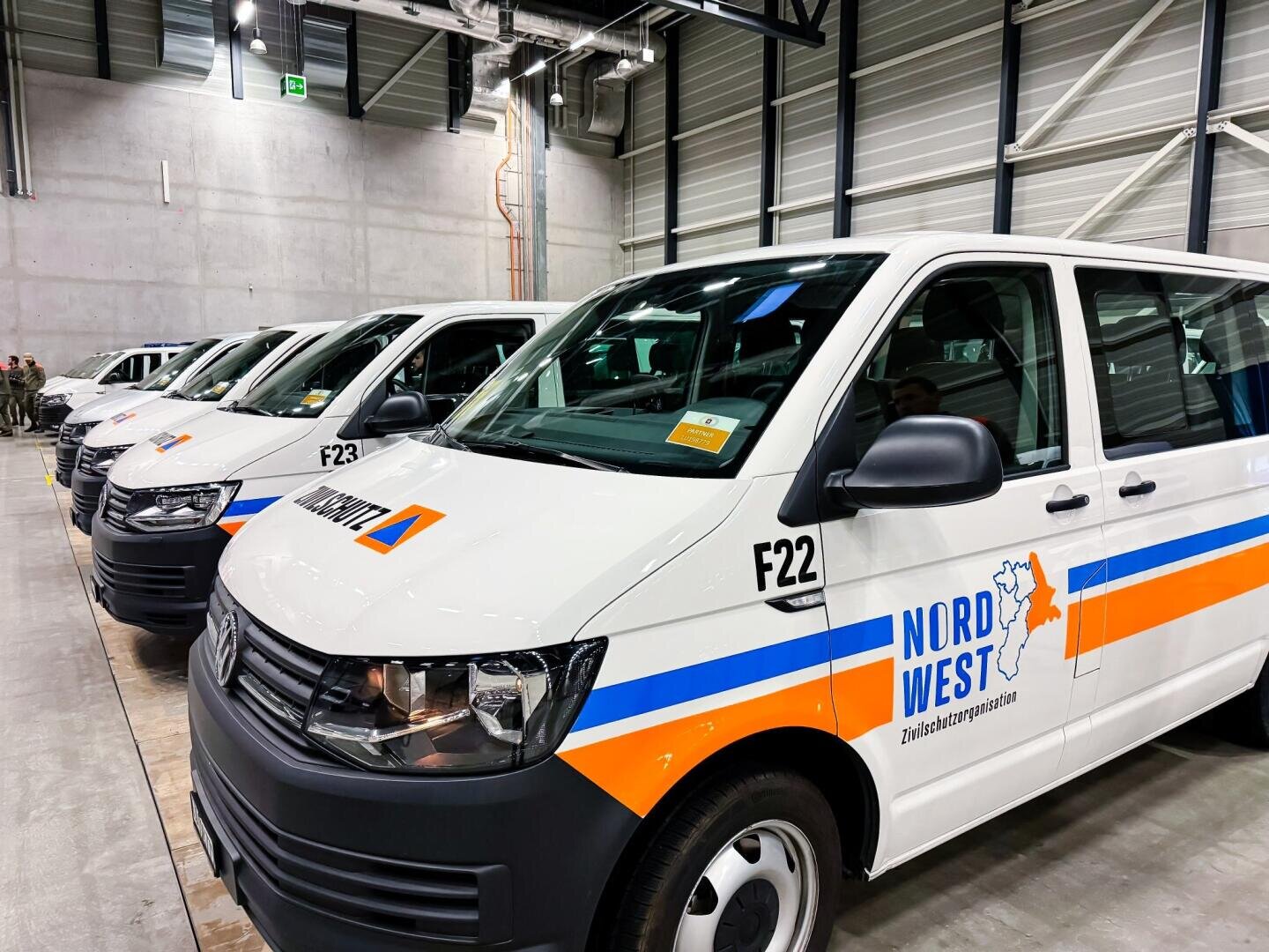 Mehrere weiße Lieferwagen mit der Aufschrift NordWest Zivilschutzorganisation und orange-blauen Streifen stehen in einer Reihe in einer großen Garage mit Betonwänden und hohen Decken.