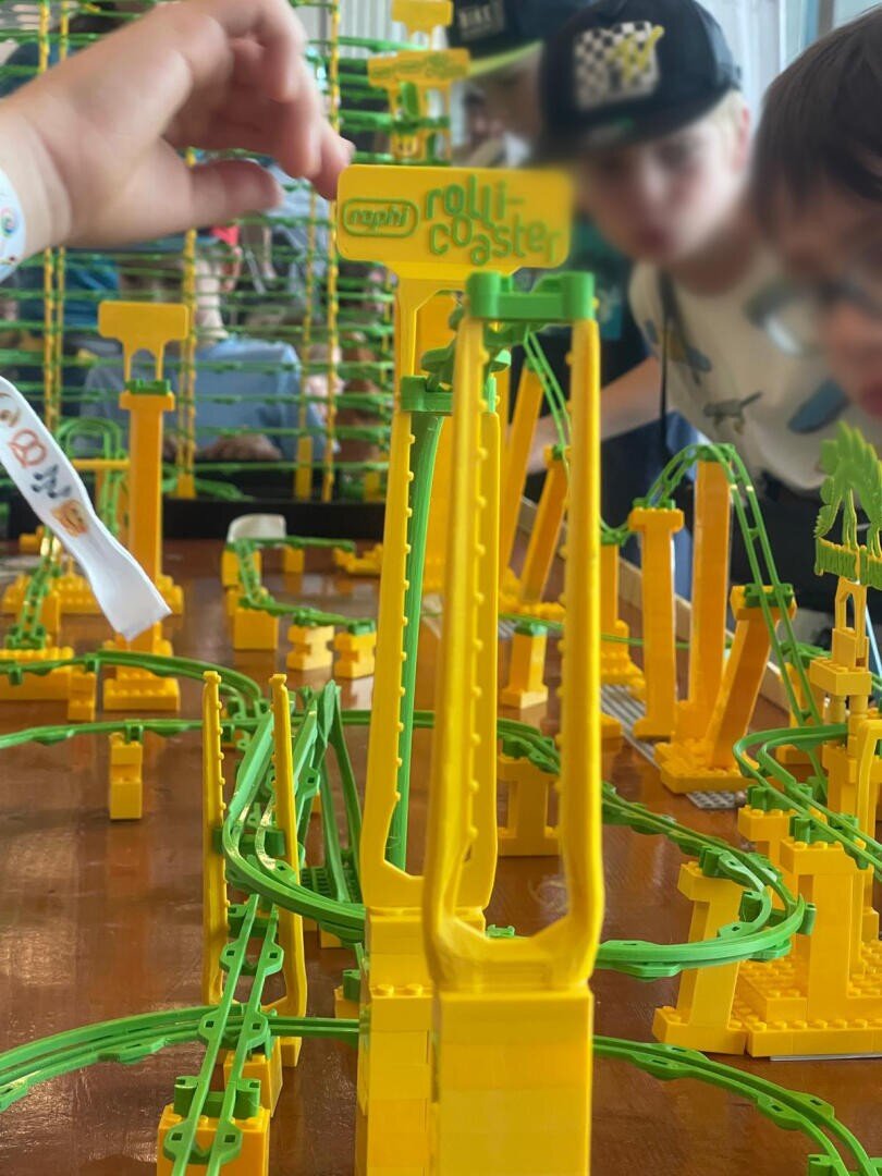Kinder spielen mit einem farbenfrohen Achterbahnspielzeugset aus Kunststoff mit gelben und grünen Schienen und Strukturen, während ein Kind den Kreisel verstellt und die anderen aufmerksam zusehen.