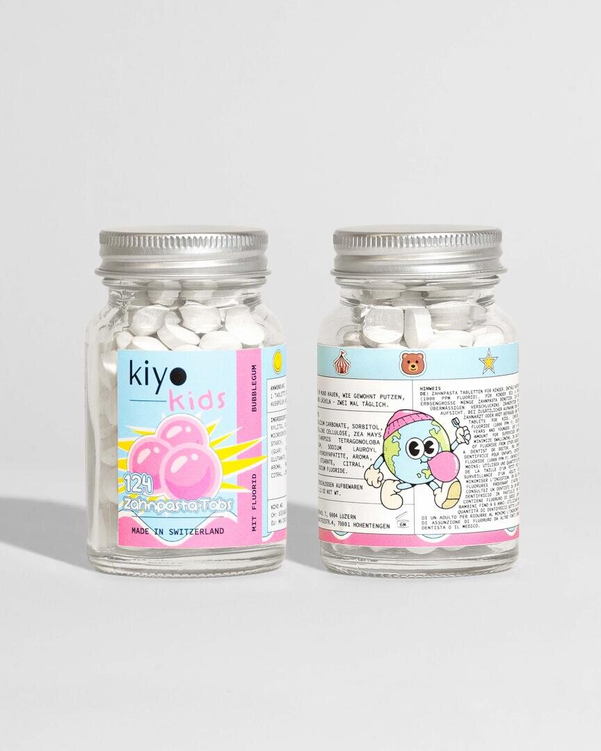 Zwei durchsichtige Gläser mit silbernen Deckeln enthalten kleine weiße Kautabletten. Die farbenfrohen Etiketten zeigen verspielte Motive, darunter eine Zeichentrickfigur und Seifenblasen, und die Aufschrift "kiyo kids 123! Zahn Pasta Tabs". Die Gläser sind in mehreren Sprachen beschriftet.