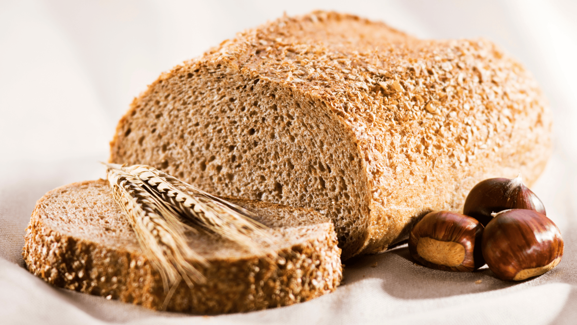 Ein Laib Vollkornbrot, in zwei Scheiben geschnitten, neben Weizenhalmen und drei braunen Kastanien auf einer hellen Unterlage platziert.