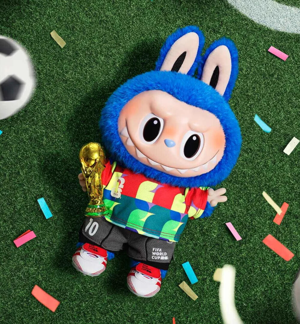 Ein Plüschtier mit einem breiten Grinsen, das einen blauen Bunny-Kapuzenpulli, ein buntes Hemd und "10 FIFA World Cup"-Shorts trägt, hält eine goldene Trophäe in der Hand, während es auf dem grünen Rasen liegt, umgeben von Konfetti und einem Fußball.