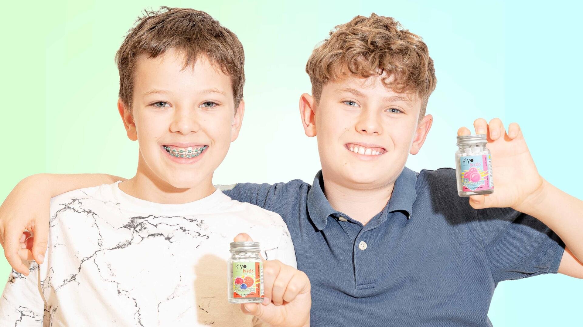 Zwei lächelnde Jungen stehen Seite an Seite und halten jeweils ein kleines Glas mit bunten Etiketten in der Hand. Ein Junge hat eine Zahnspange und trägt ein weiß marmoriertes Hemd, der andere hat lockiges Haar und trägt ein blaues Poloshirt. Der Hintergrund ist ein sanfter Farbverlauf aus Blau und Grün.