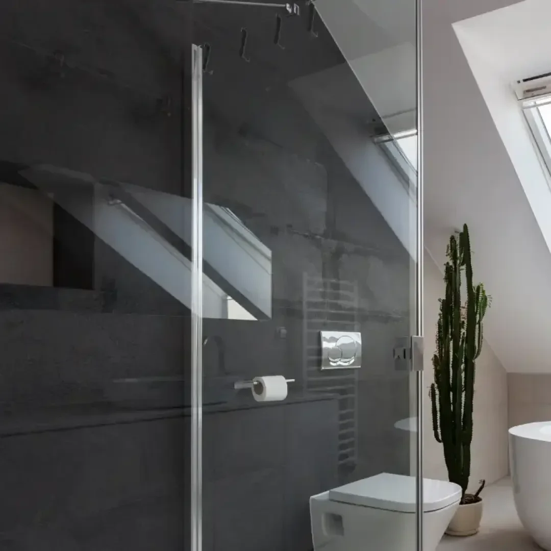 Modernes Badezimmer mit dunkelgrau gefliesten Wänden, einer gläsernen Duschabtrennung, einer weißen Toilette und einer hohen Kaktuspflanze in einem Topf in der Nähe eines Fensters in der schrägen Decke. Durch das Oberlicht fällt natürliches Licht ein.
