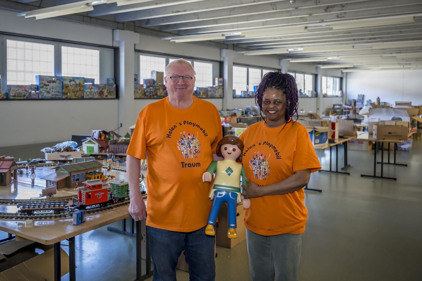 Zwei Personen, die passende orangefarbene "Heidi's Playground"-Shirts tragen, stehen in einem Raum voller Spielzeug und halten eine große Playmobil-Puppe. Auf den Tischen um sie herum sind Modelleisenbahnen und Spielzeughäuser ausgestellt.