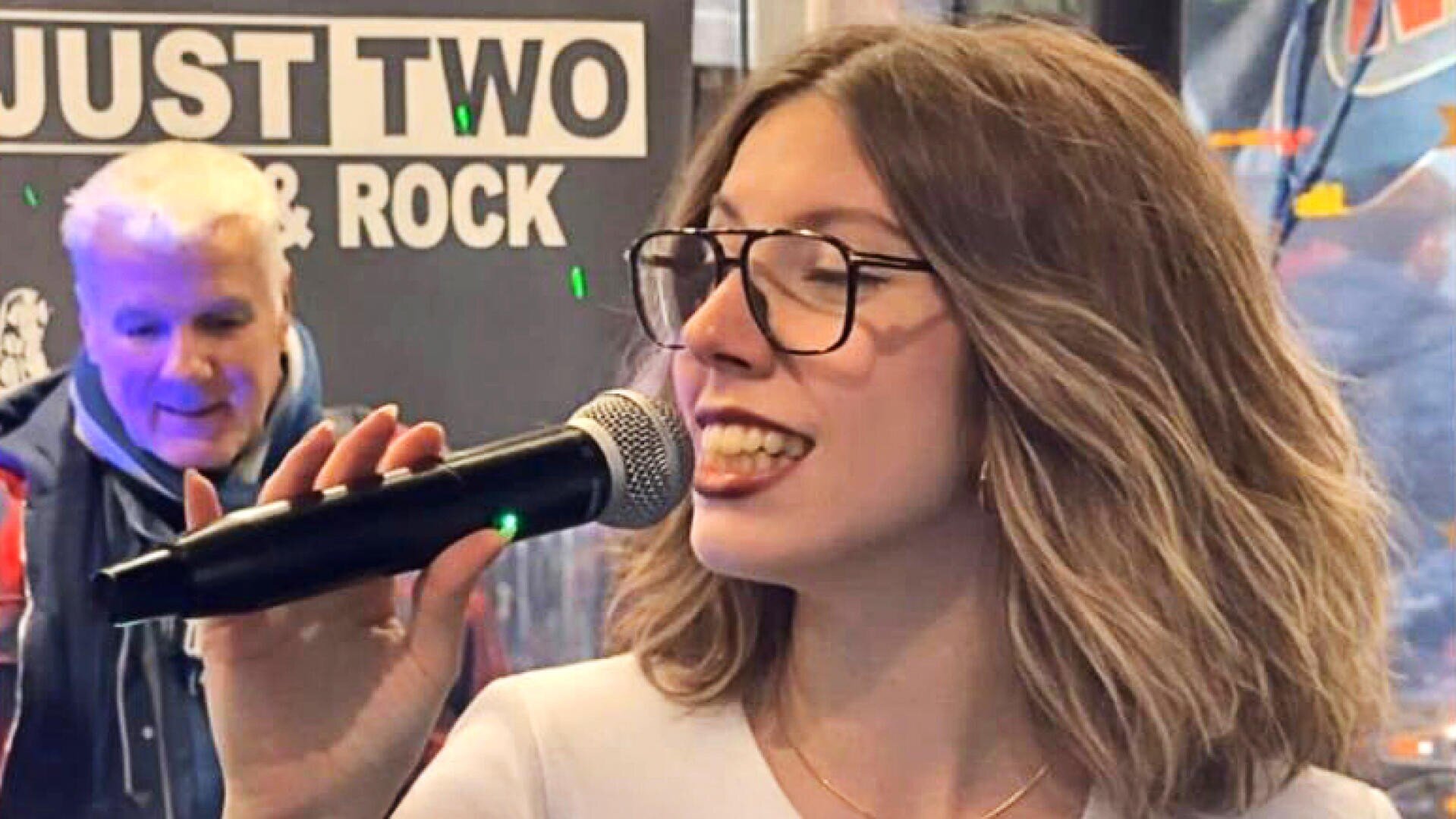 Eine Frau mit schulterlangem Haar und Brille singt in ein Mikrofon. Ein Mann steht im Hintergrund neben einem Schild mit der Aufschrift JUST TWO & ROCK. Die Szene ist lebhaft und gut beleuchtet.