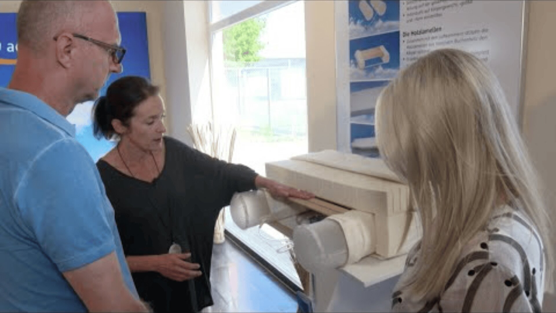 Eine Frau erklärt einem Mann und einer Frau in einem Innenraum ein weißes mechanisches Modell. Die Gruppe konzentriert sich auf das Objekt, das ein technischer oder architektonischer Prototyp zu sein scheint, in einem gut beleuchteten modernen Raum.