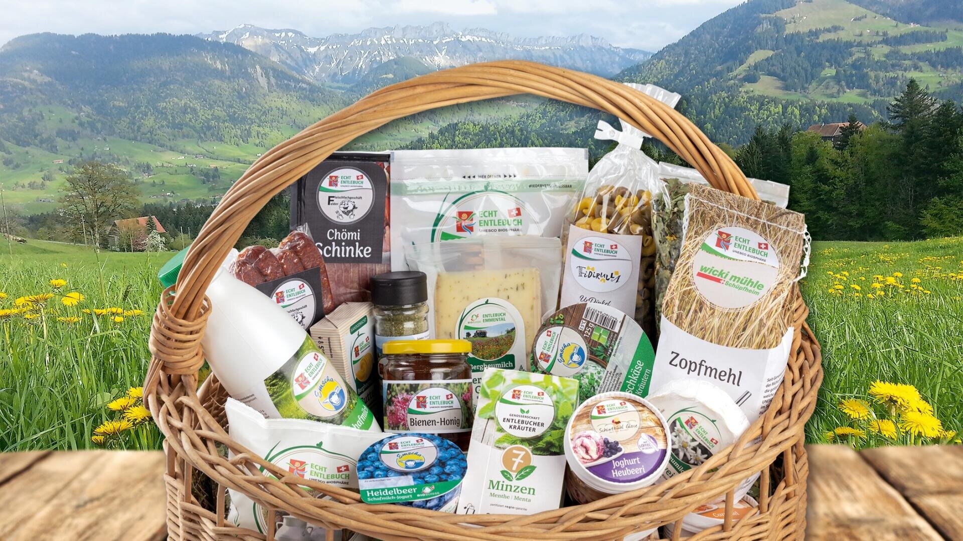 Ein Weidenkorb, gefüllt mit verschiedenen verpackten Schweizer Lebensmitteln und Getränken, einschließlich Käse, Brotmischungen, Kräutern und Marmeladen, steht auf einer Holzfläche mit einer malerischen Berg- und Wiesenlandschaft im Hintergrund.