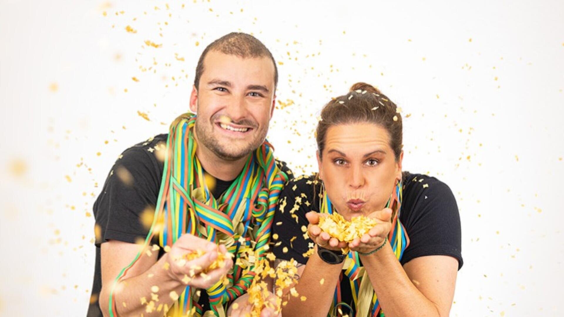 Zwei Personen, die bunte Schals und schwarze Hemden tragen, lächeln und werfen oder blasen Konfetti in die Kamera, während um sie herum vor einem weißen Hintergrund goldenes Konfetti in die Luft gestreut wird.