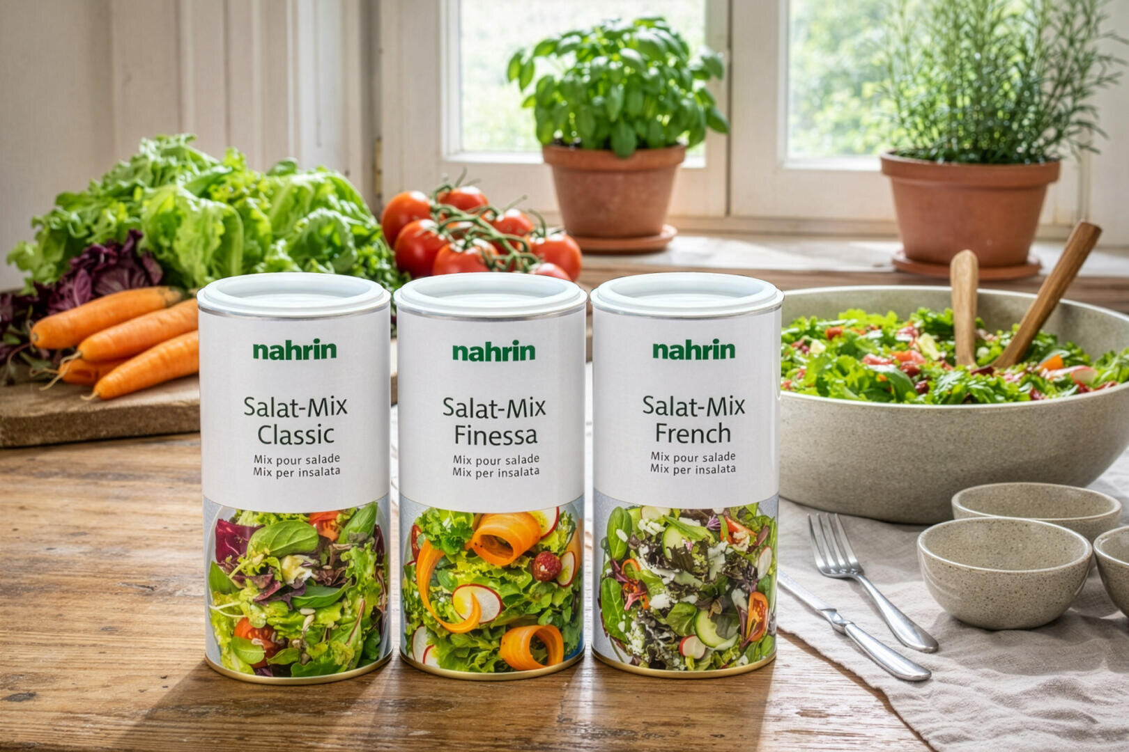 Drei Kanister mit Nahrin-Salatmischungen - Classic, Finessa und French - stehen auf einem Holztisch mit frischem Gemüse, einer Salatschüssel, Tellern, Besteck und Kräutertöpfen im Hintergrund.