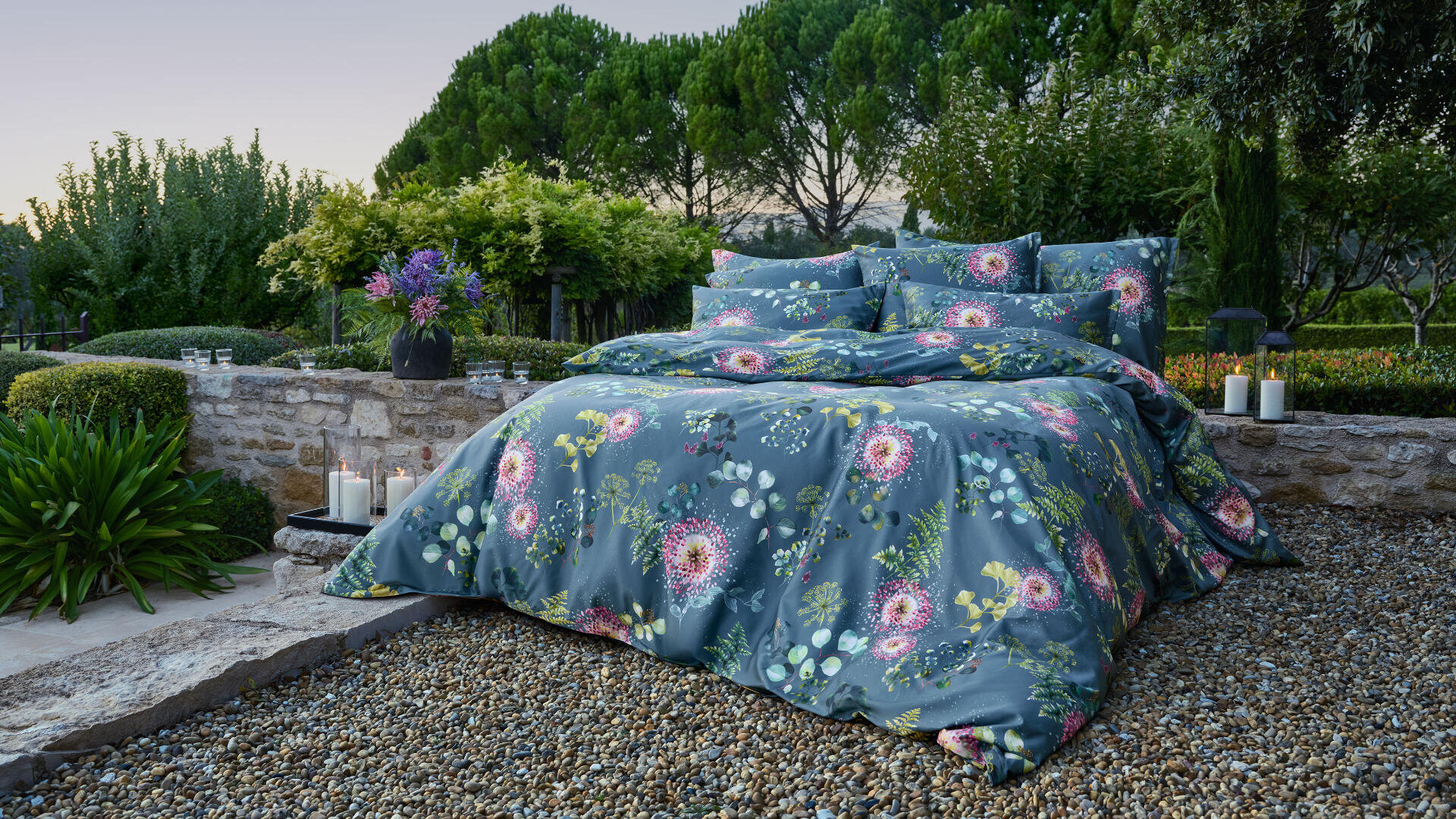 Ein Bett mit blauem, blumengemustertem Bettzeug steht im Freien auf einer Kiesfläche, umgeben von üppigem Grün, Bäumen und Steinmauern, mit Kerzen und einer Blumenvase in der Nähe.