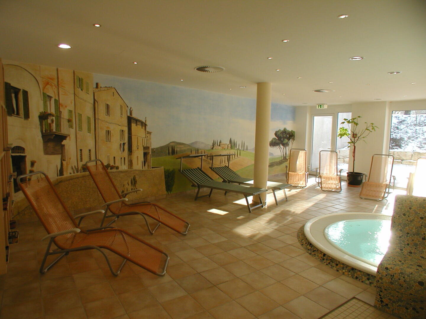 Ein Wellness-Ruheraum mit Liegestühlen, einem kleinen runden Whirlpool, gefliesten Böden und einem Wandgemälde, das eine italienische Landschaft zeigt. Durch große Fenster strömt Sonnenlicht herein, und neben dem Eingang steht eine Topfpflanze.