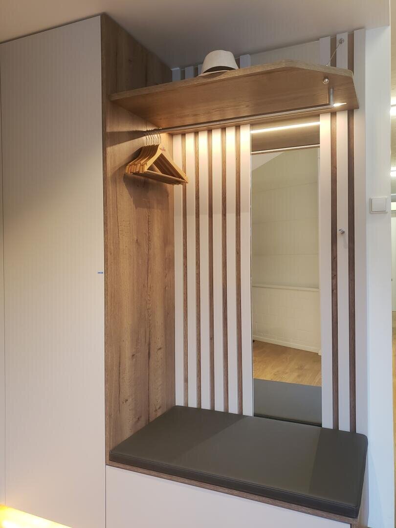 Ein moderner Eingangsbereich mit einer Holzbank, einem großen Spiegel, einem Regal mit einem Hut, mehreren Holzbügeln und vertikal gestreiften Wandpaneelen. Der Boden ist aus Holz, und in der Spiegelung ist ein Teil eines angrenzenden Raums zu sehen.