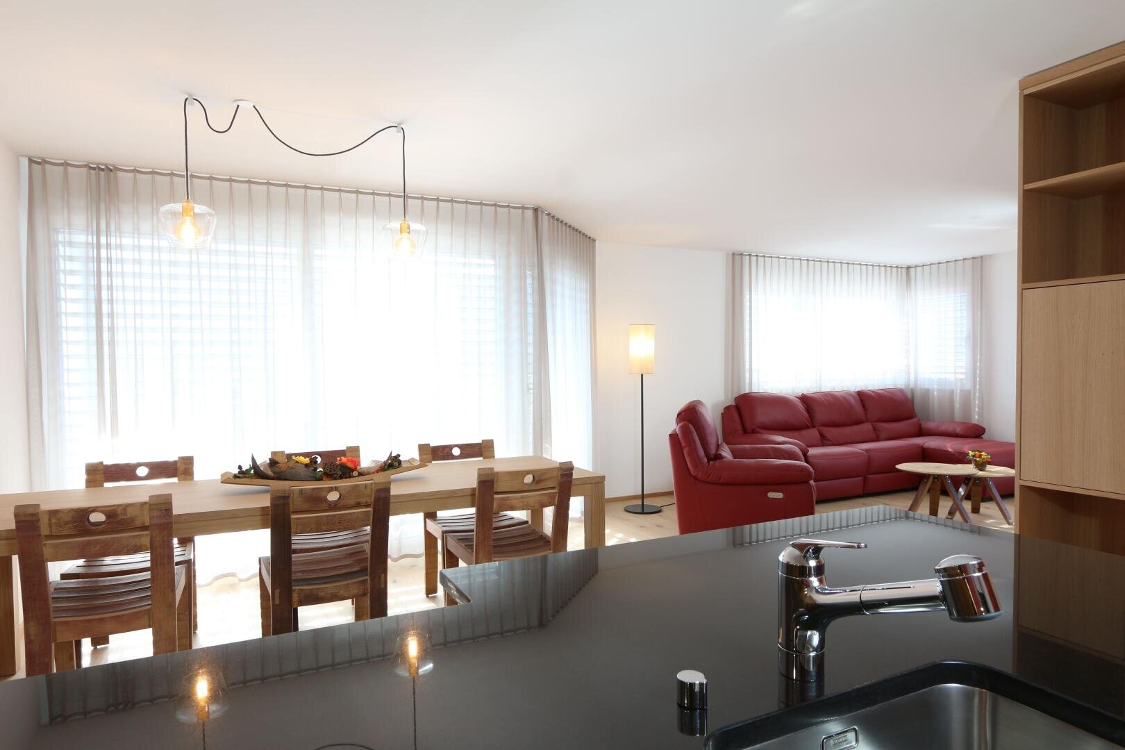 Ein moderner, heller Wohn- und Essbereich mit Holztisch und -stühlen, einem roten Sektionssofa, einer Stehlampe und großen Fenstern mit durchsichtigen Vorhängen, die von der Küchenarbeitsplatte aus betrachtet werden.