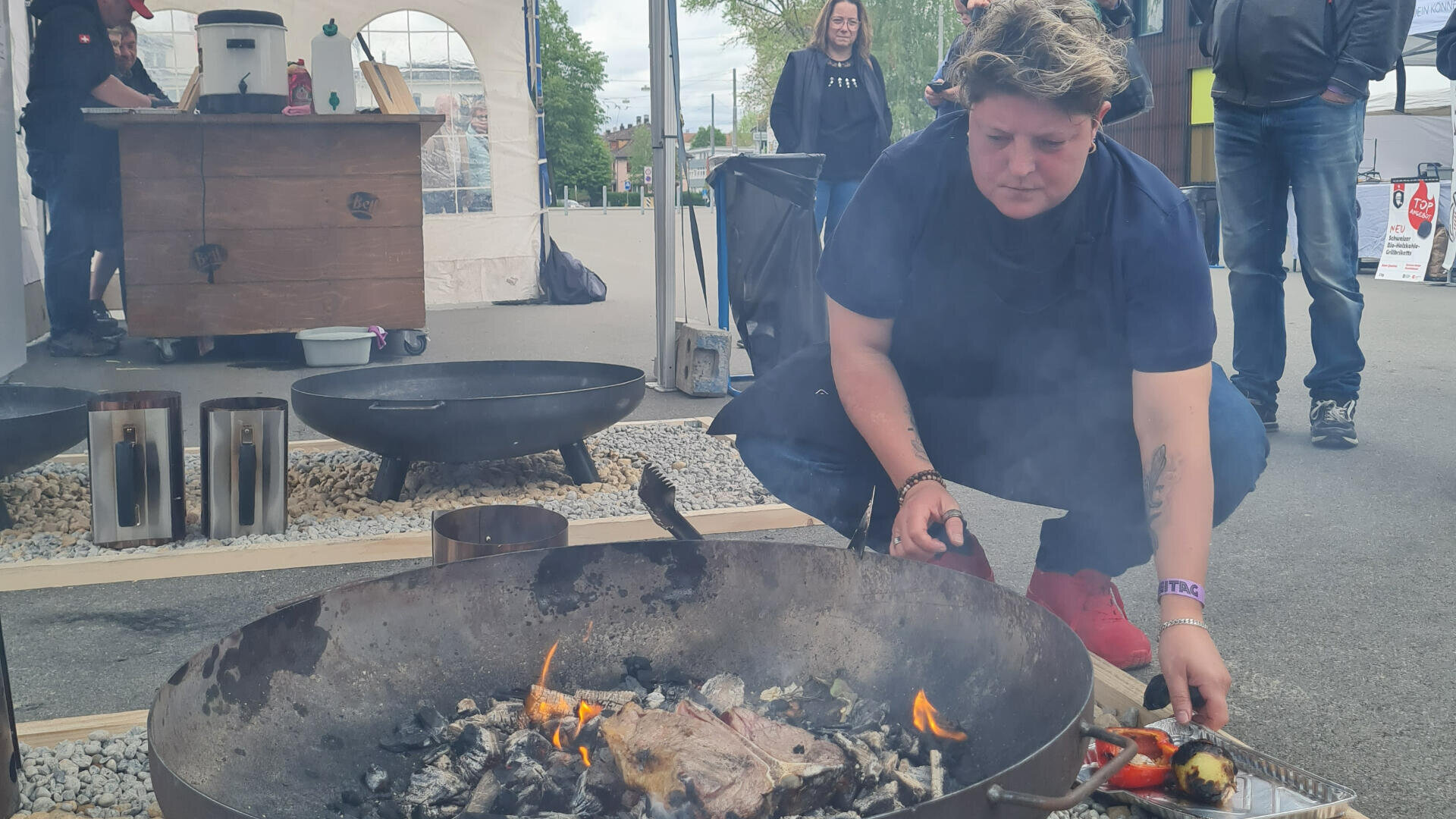 Eine Person hockt neben einer Feuerstelle im Freien und kümmert sich um Essen, das über offenen Flammen gekocht wird. Im Hintergrund sind bei bewölktem Himmel andere Menschen und Marktstände zu sehen.