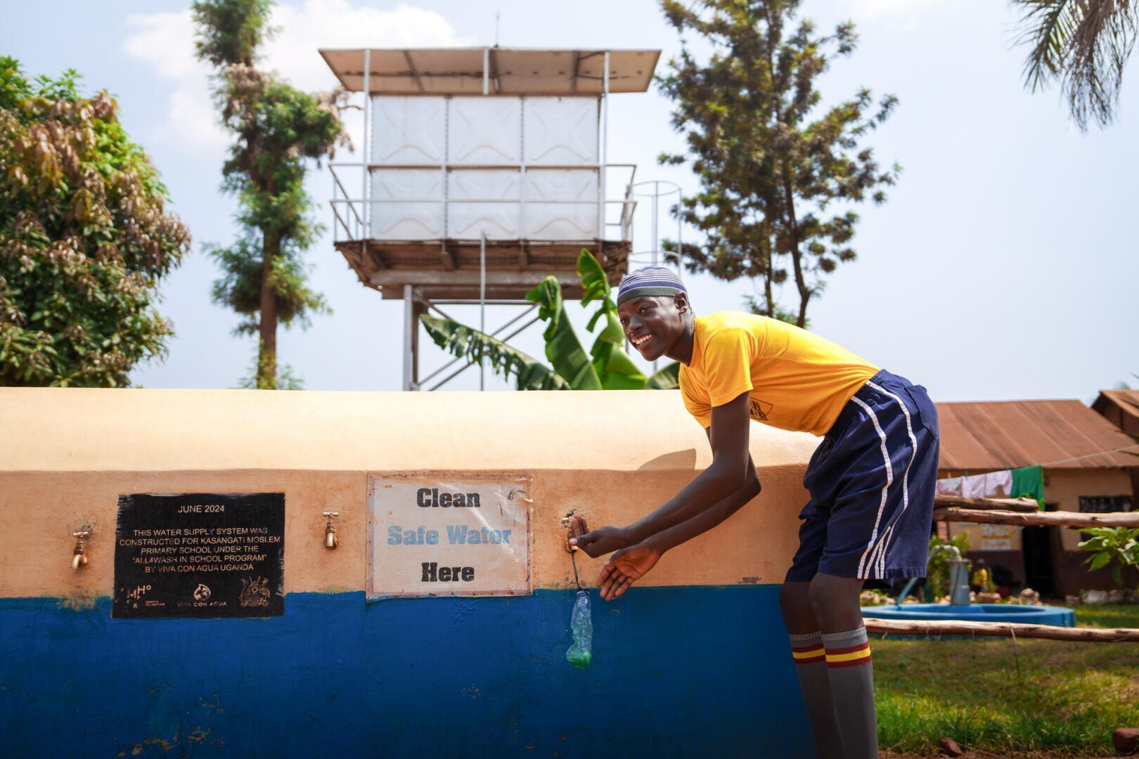 Ein lächelnder Junge in einem gelben Hemd und blauen Shorts füllt eine Flasche aus einem Wasserhahn mit der Aufschrift "Clean Safe Water Here" (Sauberes, sicheres Wasser hier), während im Hintergrund ein Wasserturm und Bäume zu sehen sind.