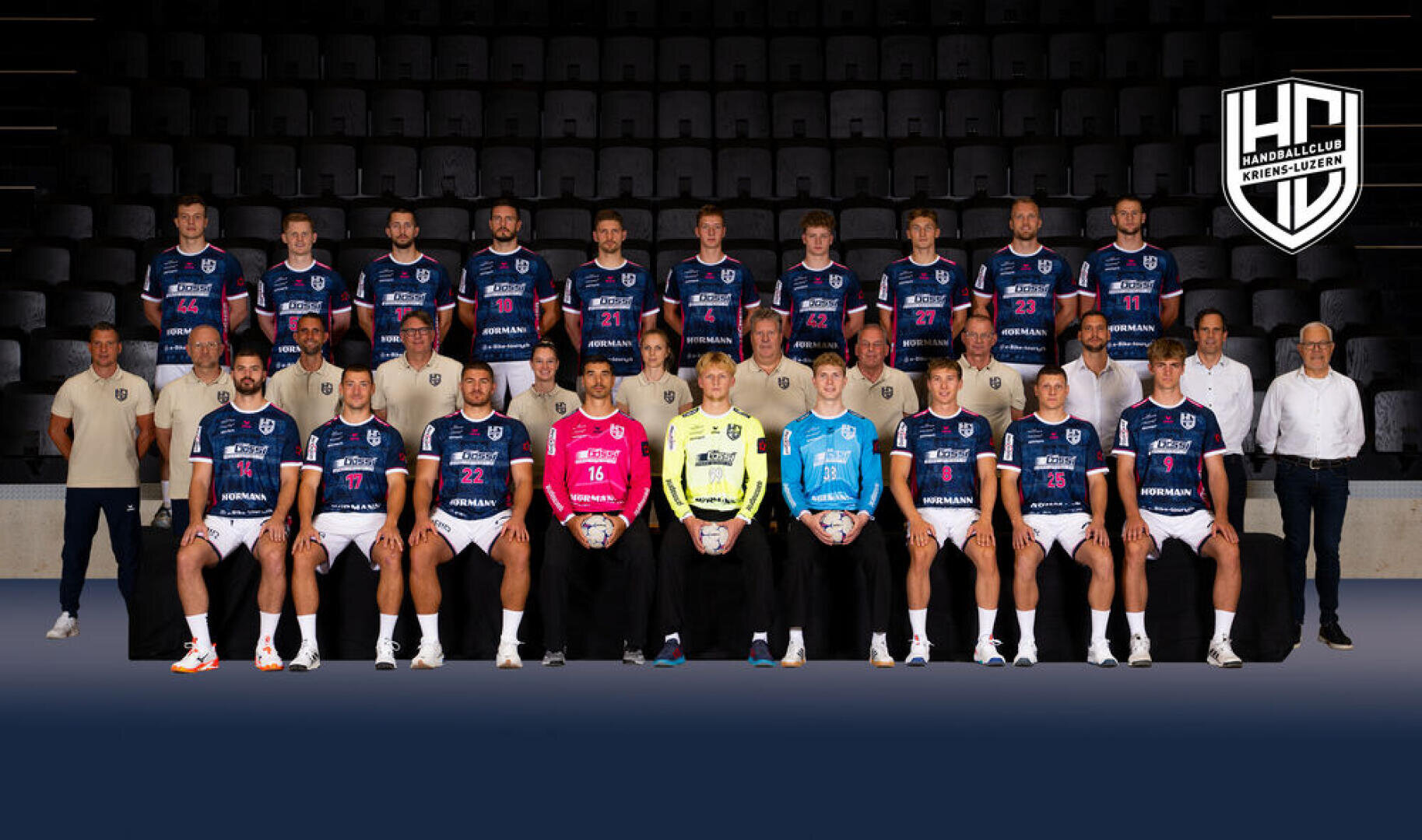 Eine Handballmannschaft und ihre Betreuer stehen in drei Reihen in einer Arena und tragen blau-weiße Trikots. Vorne sitzen drei Torhüter in verschiedenfarbigen Trikots, die Handbälle halten. In der rechten oberen Ecke ist das Logo der Mannschaft zu sehen.