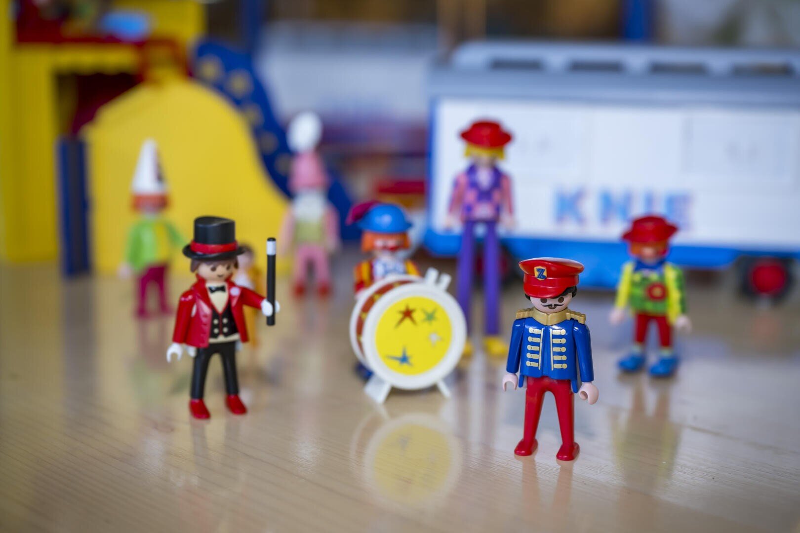 Mehrere bunte Spielzeugfiguren in Zirkuskostümen, darunter ein Zirkusdirektor und ein Trommler, stehen auf einer hölzernen Fläche mit weiteren Spielzeugfiguren und einem weißen Zirkuswagen, der im Hintergrund verschwimmt.