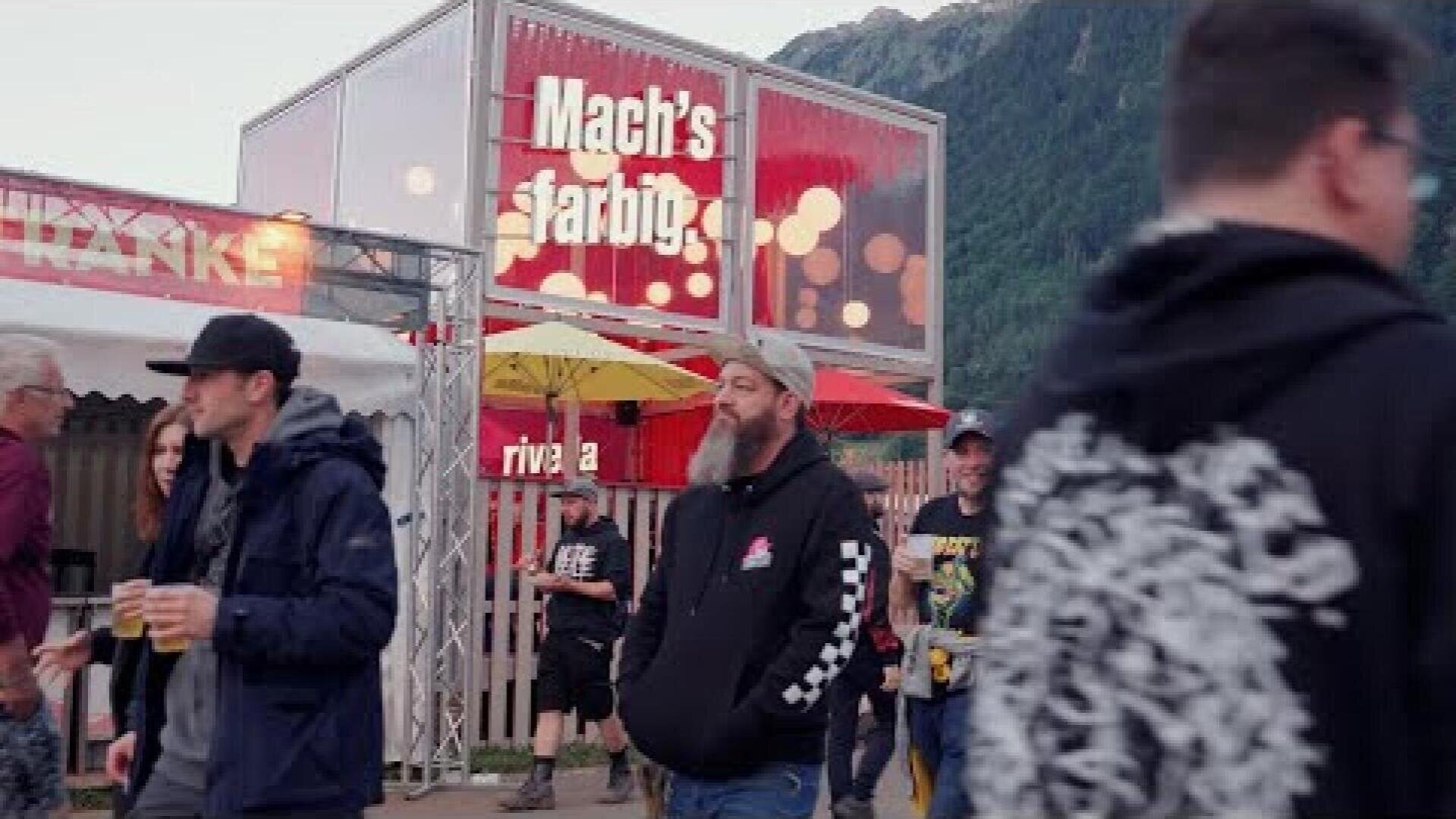 Menschen gehen bei einer Veranstaltung oder einem Festival im Freien spazieren, mit Bergen im Hintergrund. Auf einem Schild an einem Gebäude steht "Machs farbig". Einige Teilnehmer tragen Kapuzenpullis und Hüte, und eine Person hält ein Getränk in der Hand.