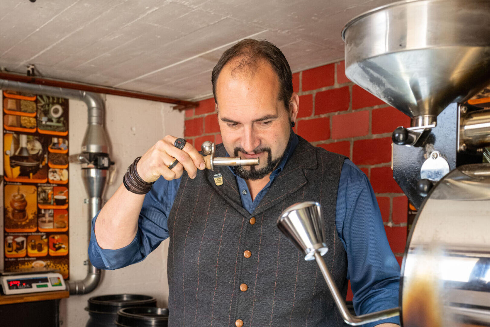 Ein Mann in blauem Hemd und Weste riecht an einem Kaffeeprobengerät neben einer großen Kaffeeröstmaschine in einem Raum mit roten Backsteinwänden und Kaffeepostern.