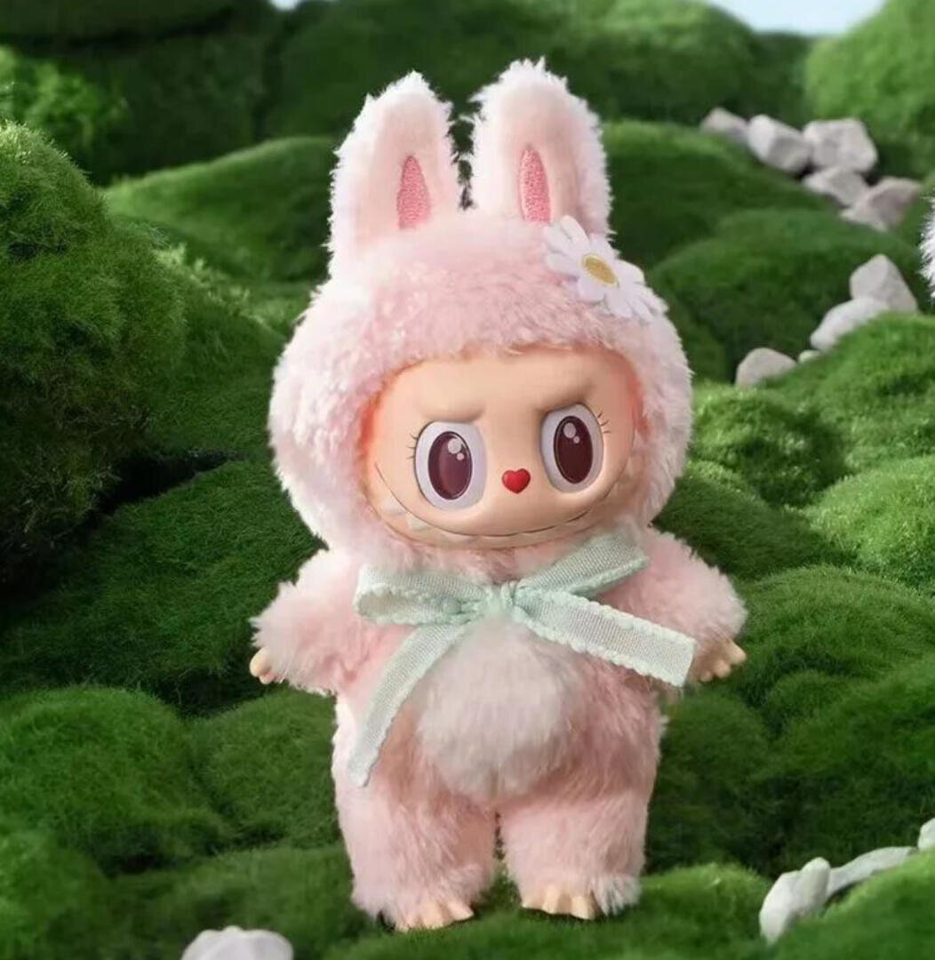 Eine niedliche Puppe mit großen Augen, einer herzförmigen Nase und einer Blume auf dem Kopf, gekleidet in ein flauschiges rosa Hasenkostüm mit grüner Schleife, steht auf saftig grünem Gras mit Felsen im Hintergrund.