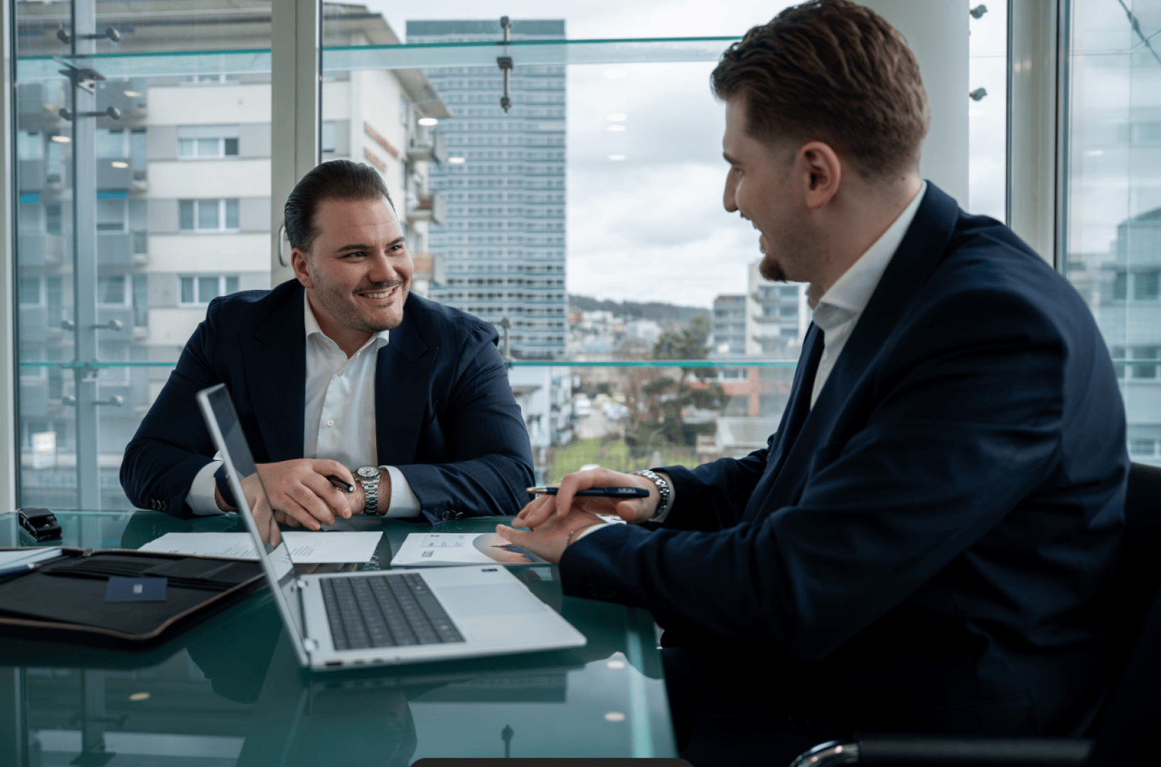 Zwei Männer in Geschäftsanzügen sitzen an einem Glastisch in einem modernen Büro, lächeln und diskutieren über Dokumente. Auf dem Tisch liegen ein Laptop und Papiere. Im Hintergrund sind große Fenster und städtische Gebäude zu sehen.