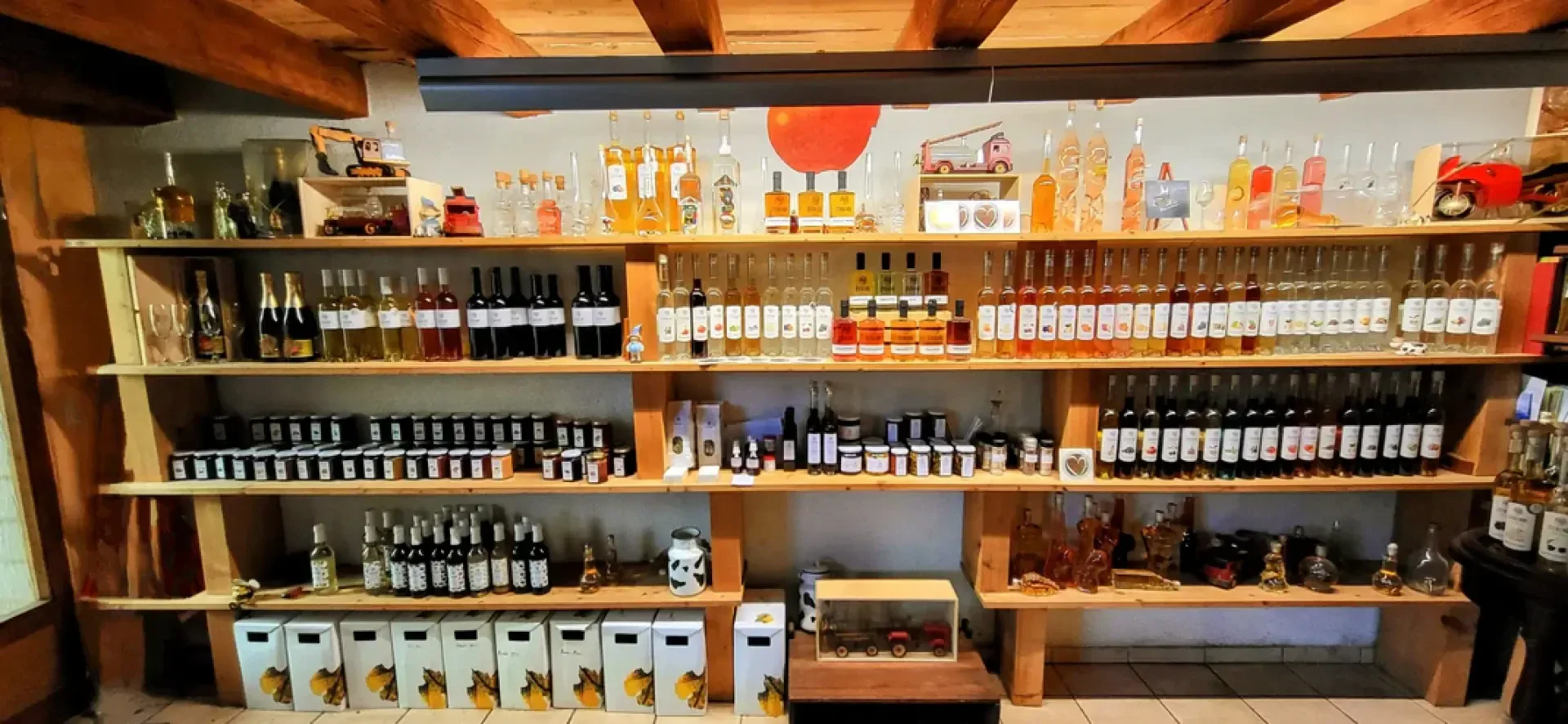 In den Holzregalen sind Wein- und Spirituosenflaschen sowie Konservengläser fein säuberlich in Reihen angeordnet. In den Regalen stehen auch Kisten, Gläser und Dekorationsartikel vor einer rustikalen Wand mit einem roten Sonnenmotiv.