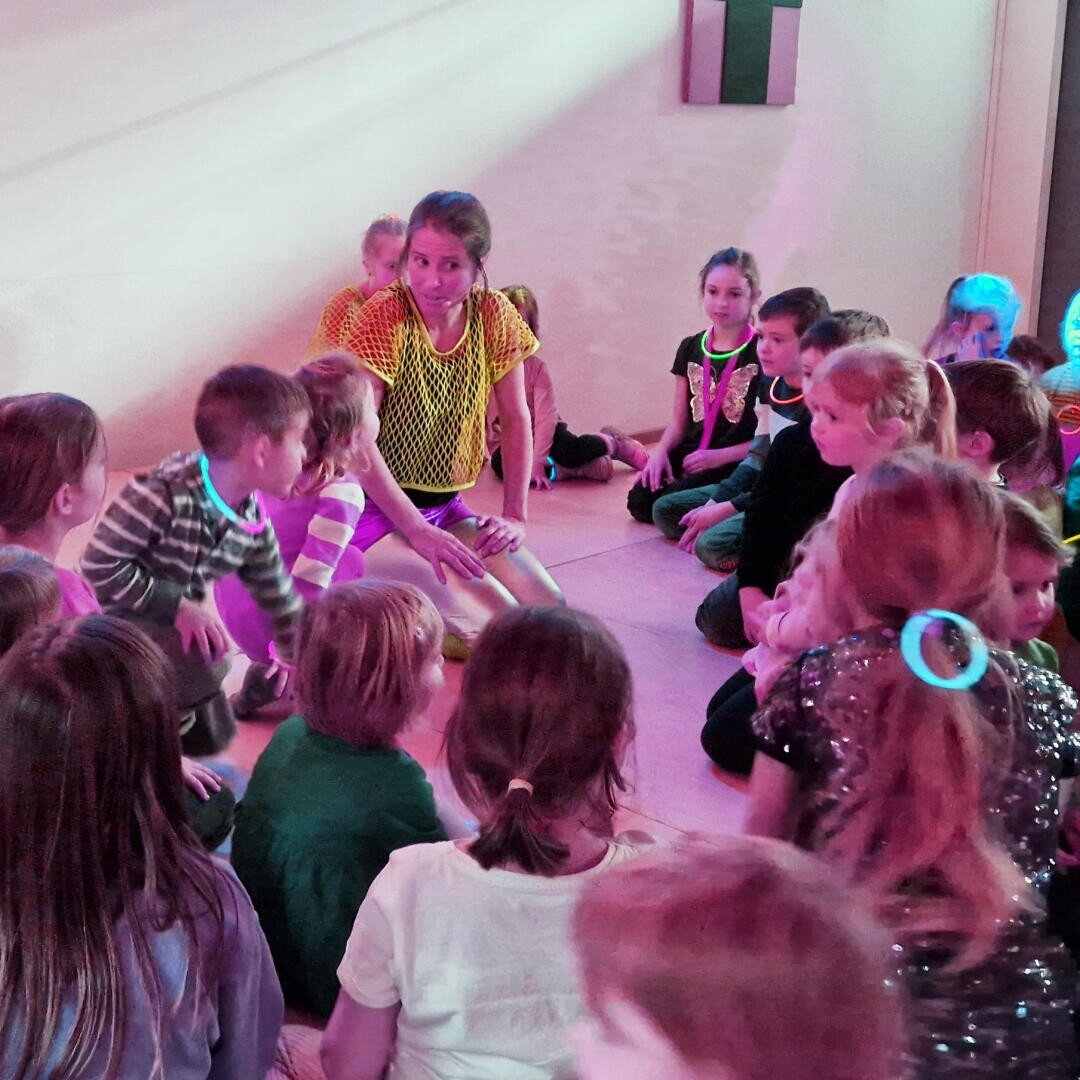 Eine Gruppe von Kindern sitzt auf dem Boden in zwei Reihen einander gegenüber und trägt bunte Kleidung und leuchtende Halsketten. Die Szene ist lebhaft, und die sanfte rosa und blaue Beleuchtung schafft eine lustige, partyähnliche Atmosphäre.