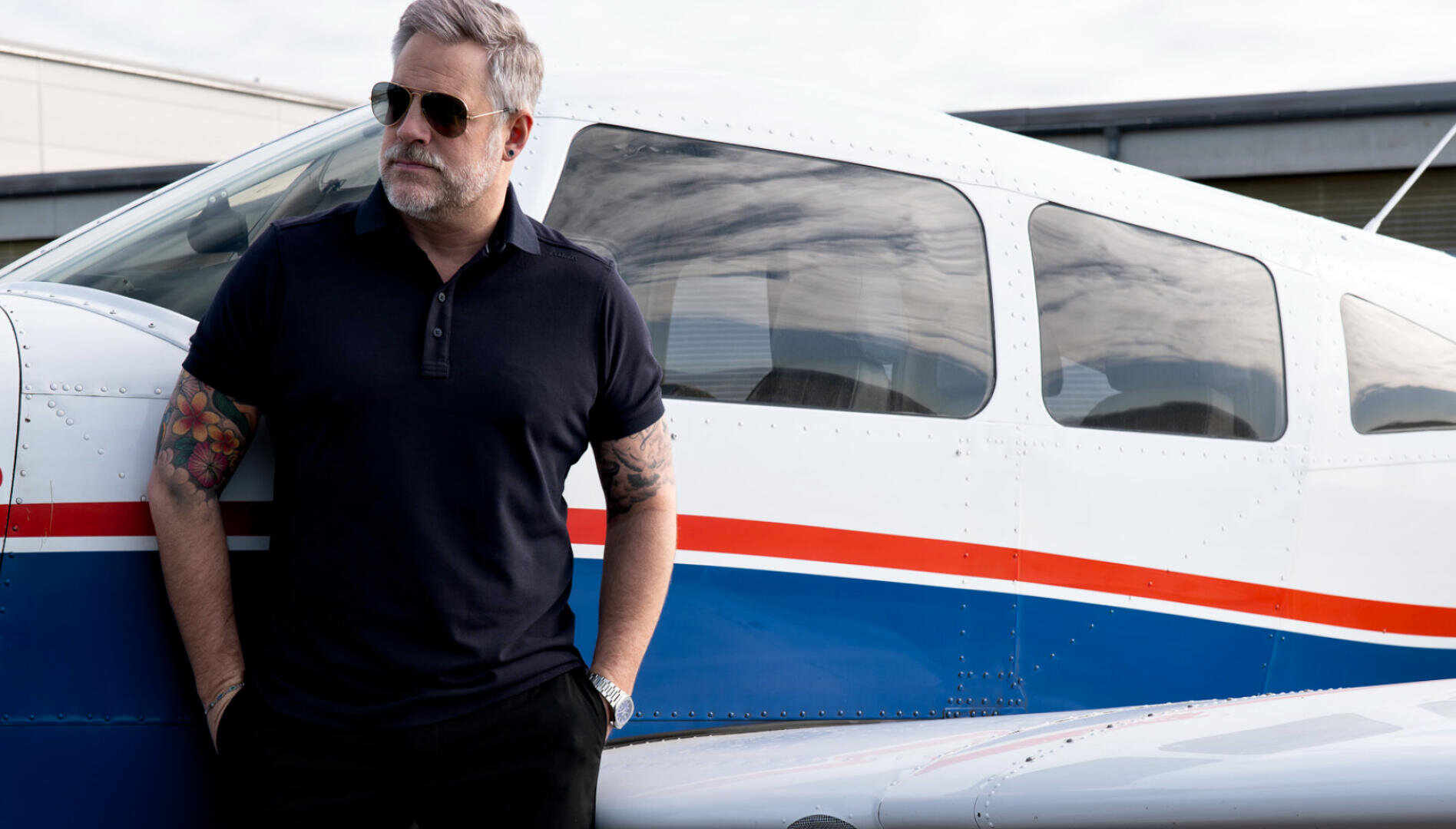 Ein Mann mit grauem Haar und Sonnenbrille, der ein schwarzes Poloshirt trägt, steht neben einem kleinen weißen Flugzeug mit blauen und roten Streifen. Er hat Tätowierungen auf seinem rechten Arm und steckt die Hände in die Taschen.